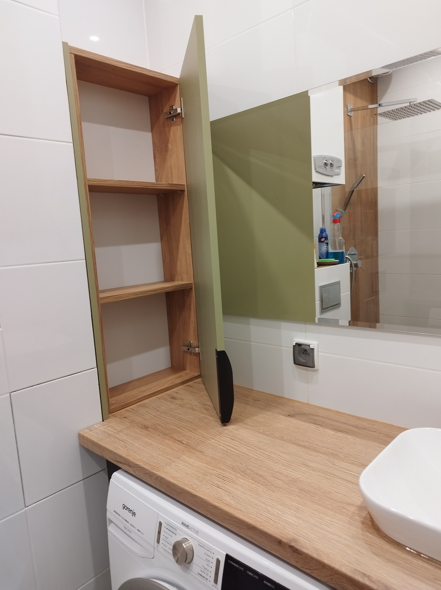 Łazienka z szafką z otwartymi półkami i zielonymi drzwiczkami, blat z drewnopodobnego laminatu nad pralką, umywalka w rogu.
