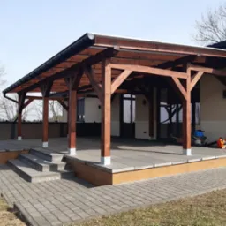 Zadaszenie tarasu z drewnianą konstrukcją, wsparte na słupach z metalowymi podstawami, z widocznymi schodami wejściowymi i elementami wykończeniowymi z imitacji drewna.