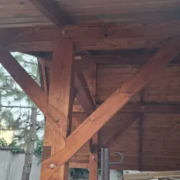 Drewniana konstrukcja z belek i słupów, widok od dołu na zadaszenie, elementy łączone drewnianymi kołkami, w tle zieleń ogrodu i budynek.