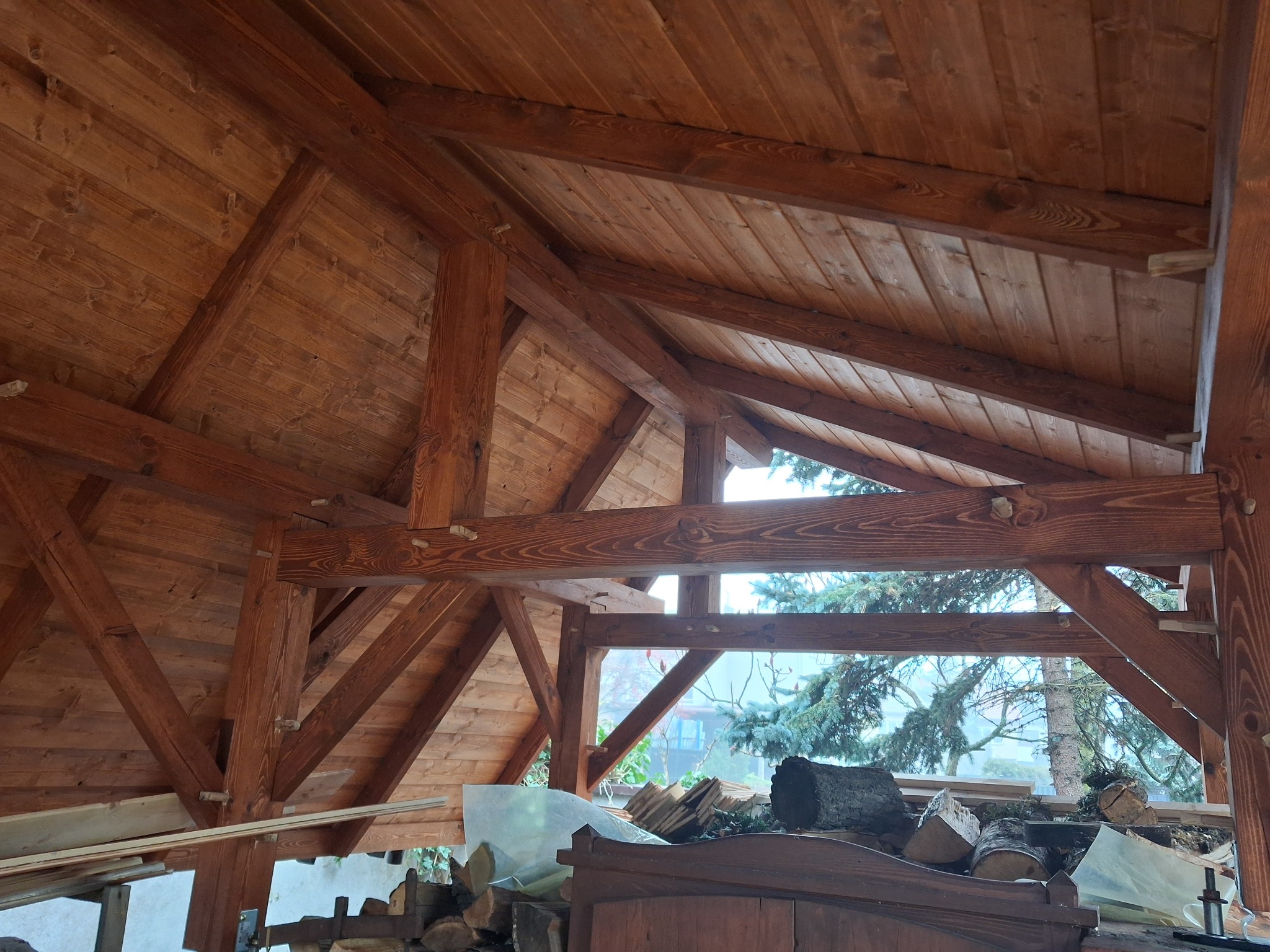 Wnętrze drewnianej konstrukcji z widoczną więźbą dachową i fragmentem otoczenia zewnętrznego przez otwór okienny, na pierwszym planie składowane drewno.