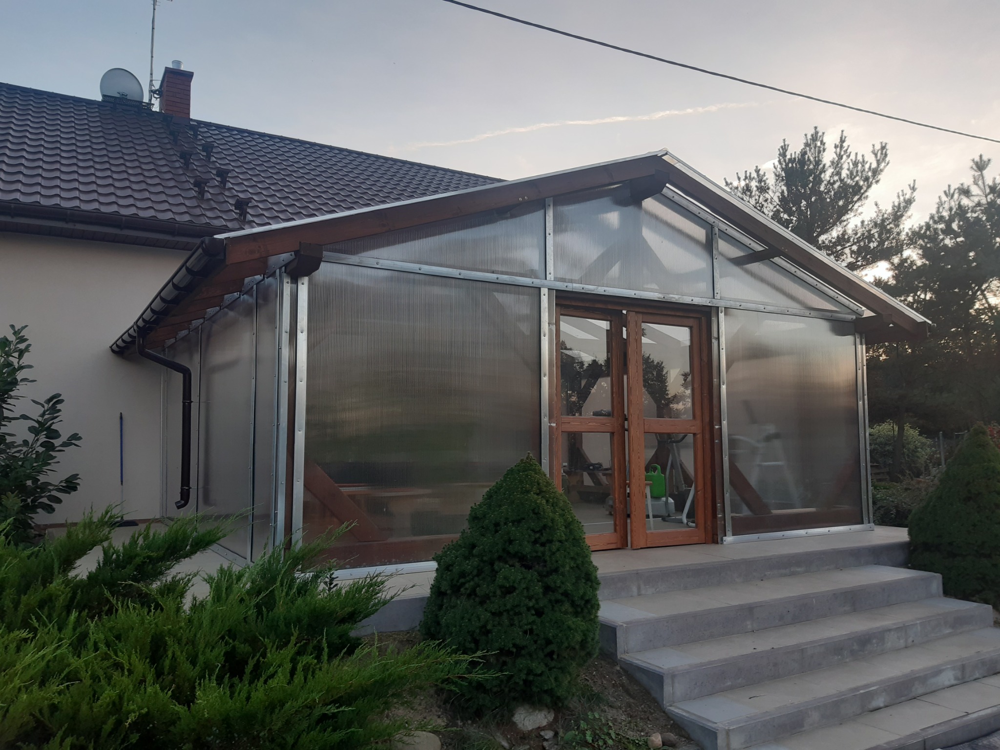 Zadaszone wejście do domu z przezroczystymi ścianami i drewnianą ramą, schody wejściowe z szarej kostki, widok z zewnątrz w pochmurny dzień.