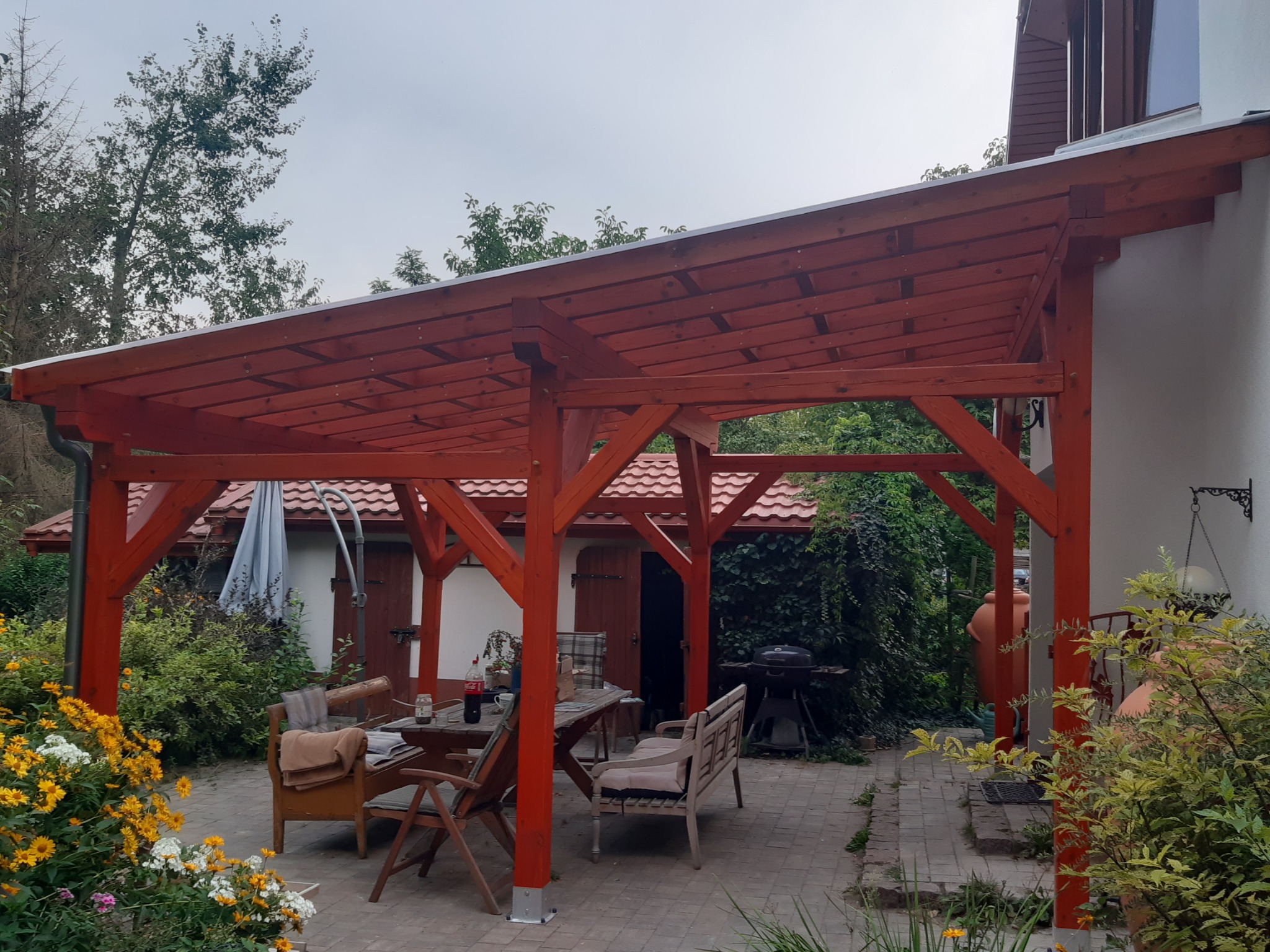 Zadaszona pergola z drewna w kolorze terakoty, z metalowym dachem, pod którą znajduje się stół z krzesłami i ławką, a w tle widoczny grill i budynek gospodarczy