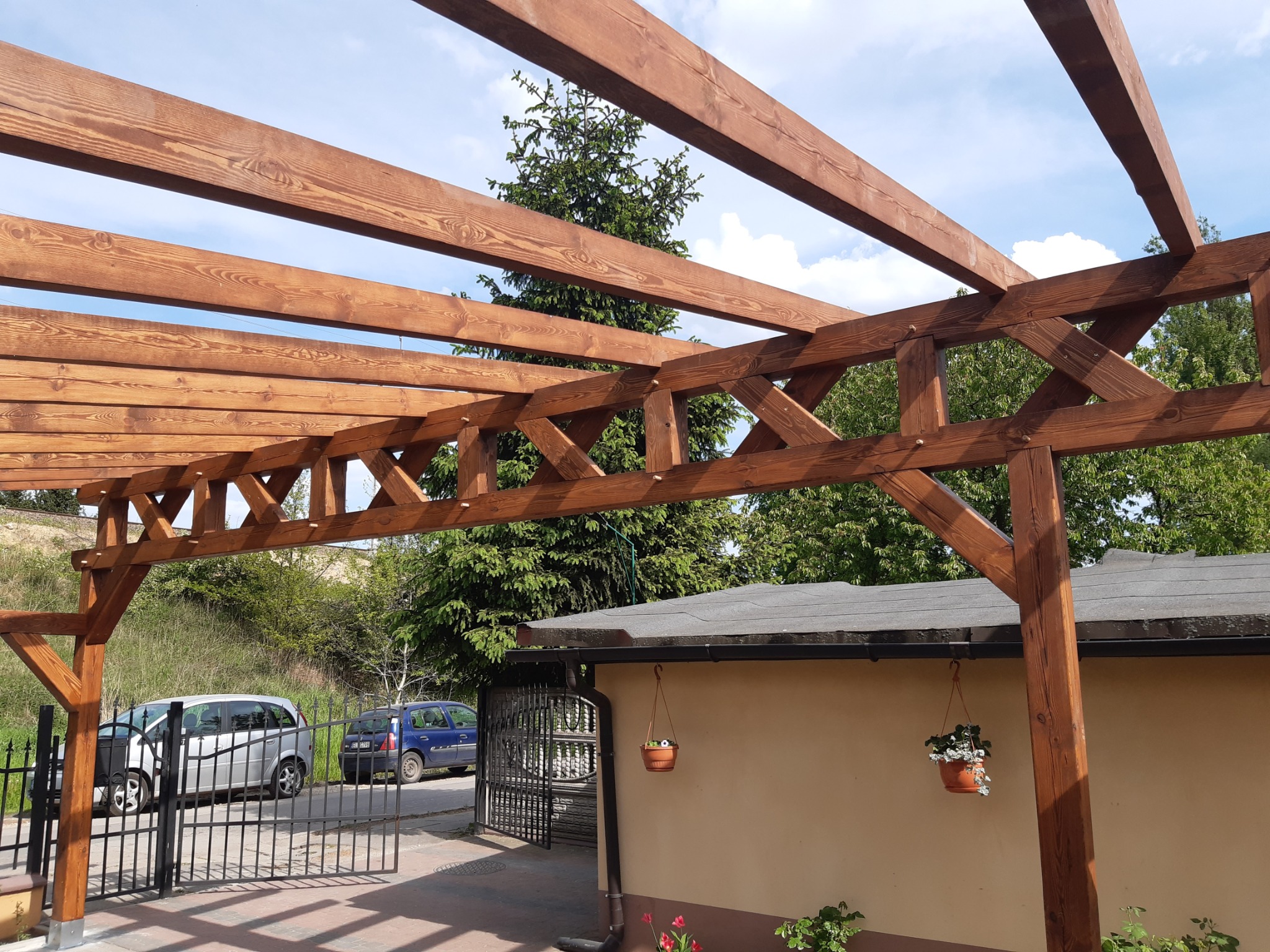 Konstrukcja z ciemnego drewna, prawdopodobnie zadaszenie, widok z dołu na niebo, z tyłu budynek gospodarczy z wiszącymi doniczkami i samochody osobowe zaparkowane na zewnątrz.
