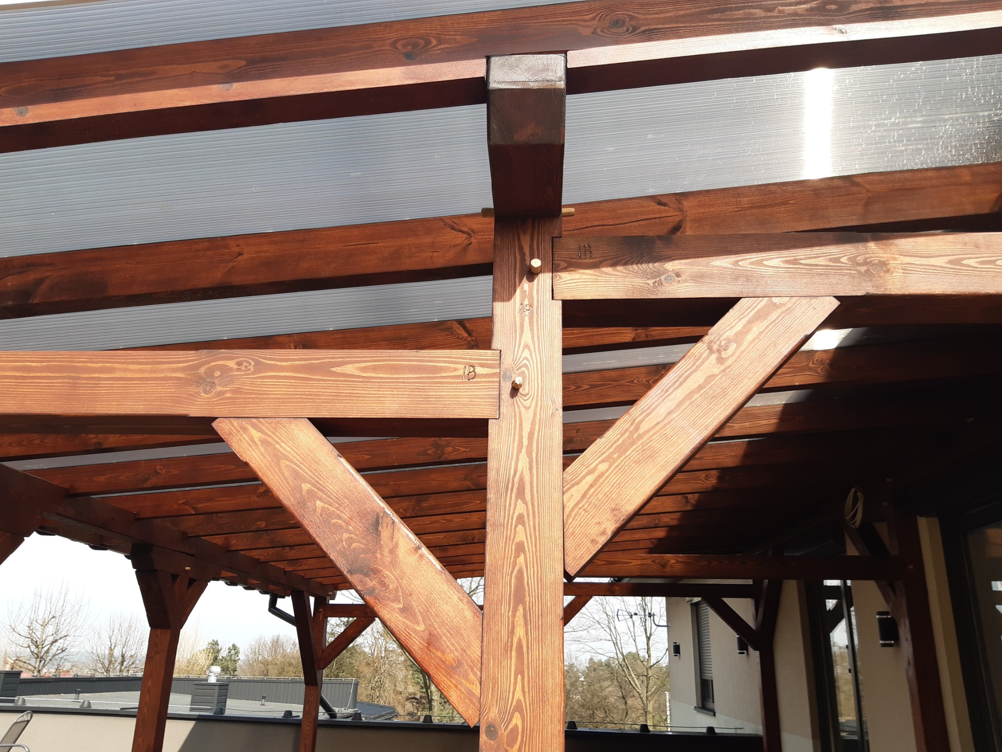 Konstrukcja z ciemnego drewna, prawdopodobnie zadaszenie tarasu lub pergola, ze sztywnym, przezroczystym dachem z tworzywa sztucznego, widoczne elementy mocujące i fragment otoczenia.