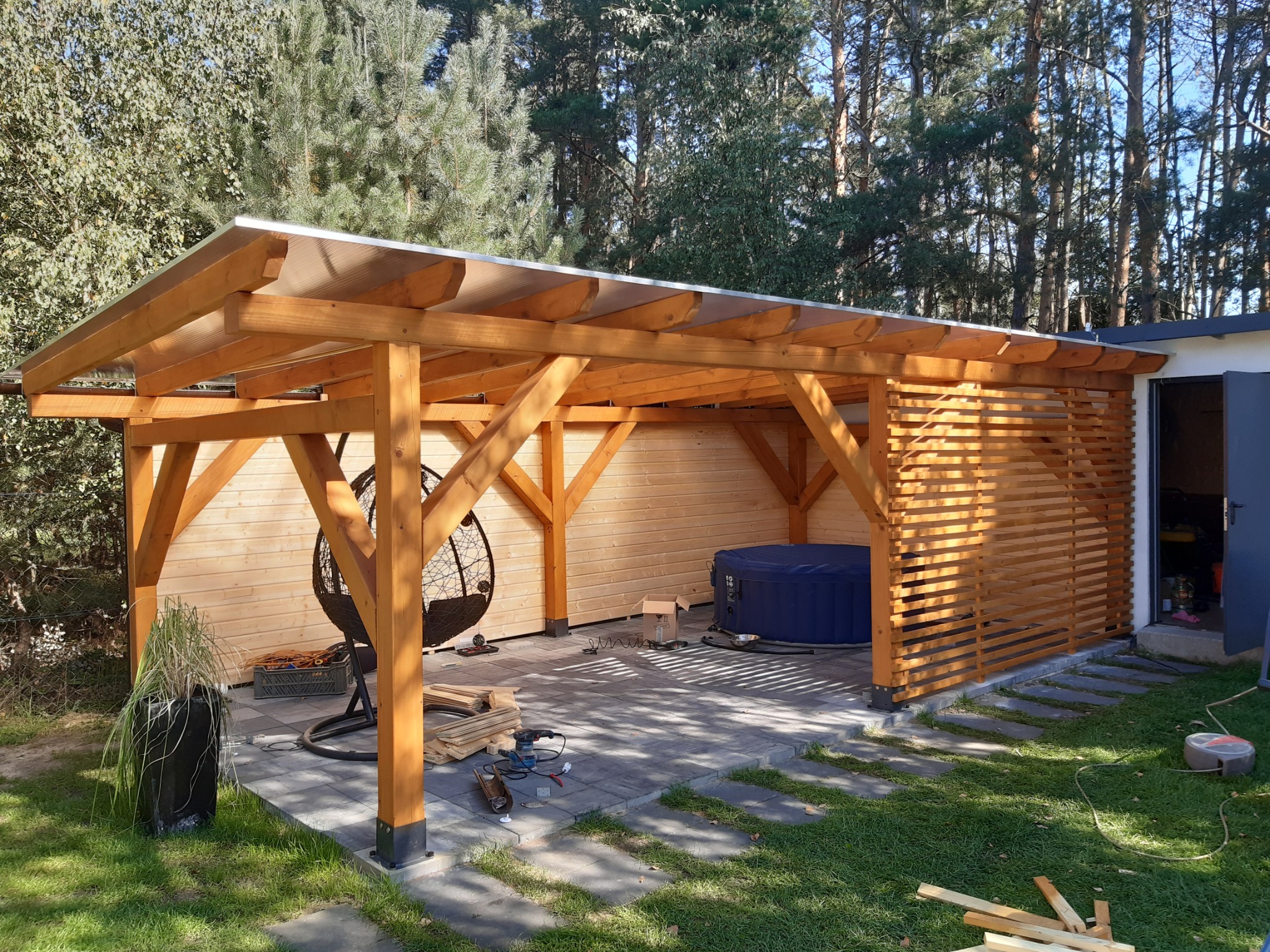 Drewniana konstrukcja z przezroczystym zadaszeniem, częściowo osłonięta drewnianymi lamelami, z jacuzzi i wiszącym fotelem w środku, na zewnątrz widoczne narzędzia i materiały budowlane.