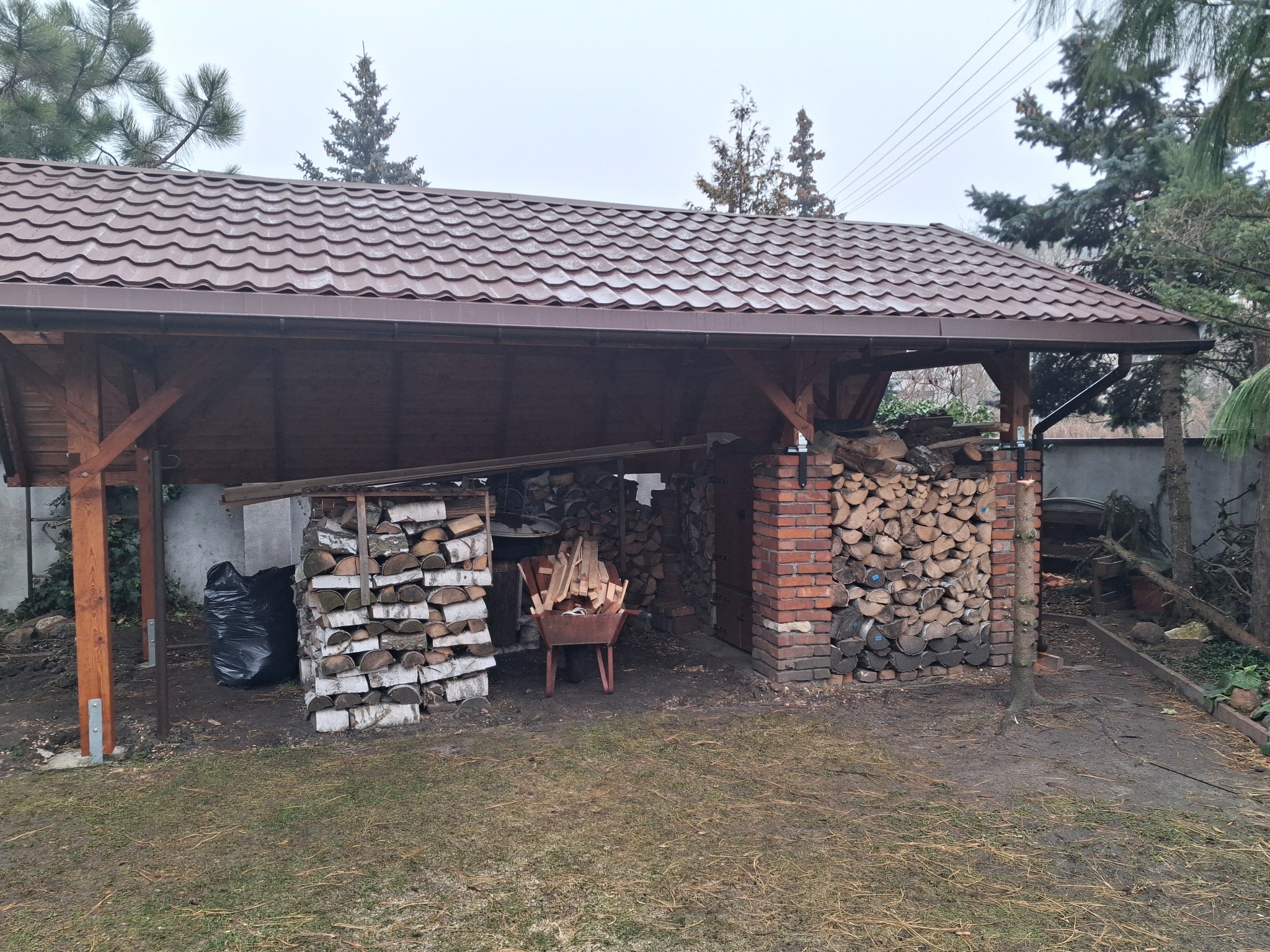 Zadaszona wiata z drewnianymi słupami i brązowym dachem z blachodachówki, służąca jako skład drewna opałowego, z widocznymi stosami drewna i taczką wypełnioną szczapami.