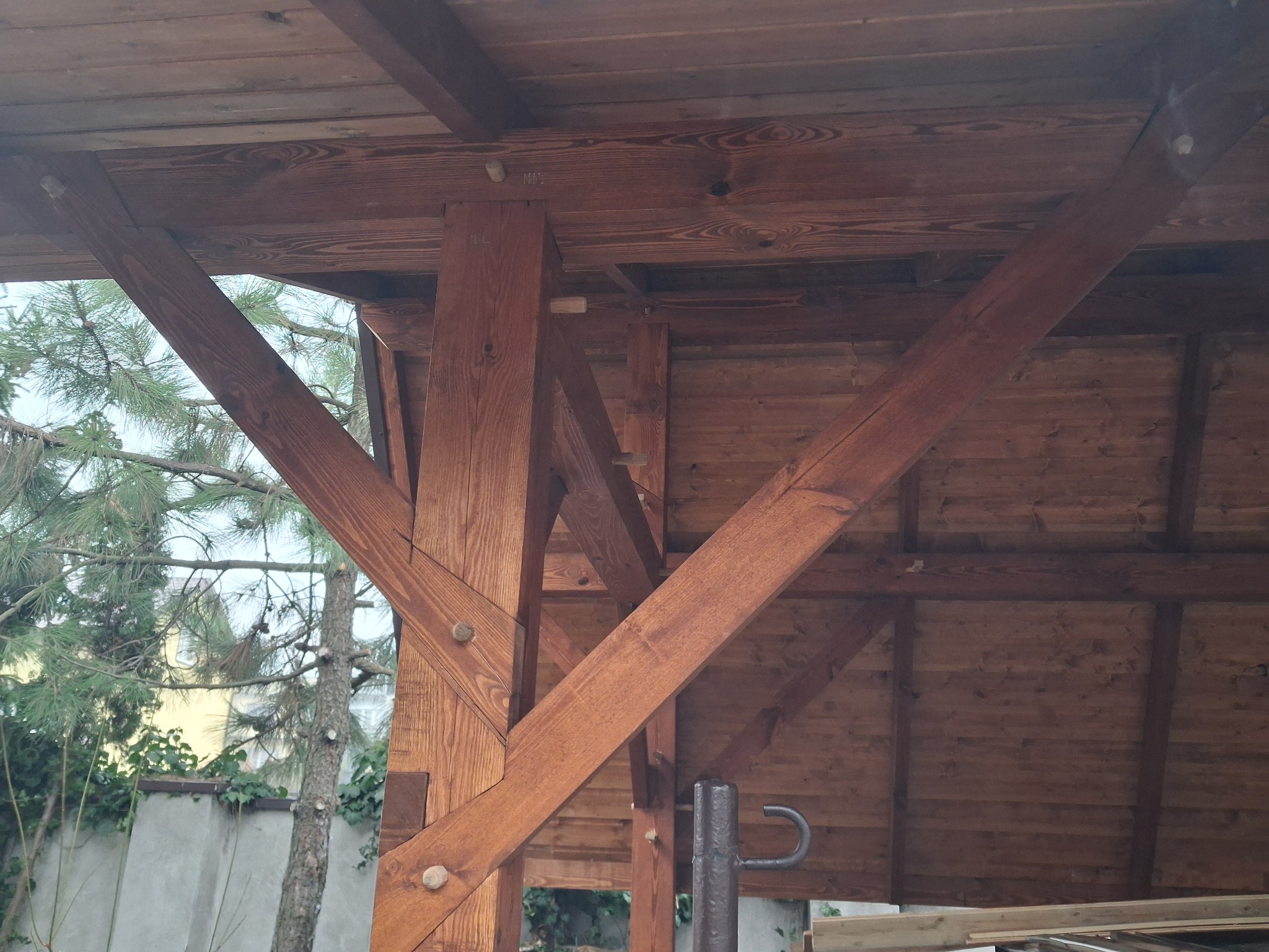 Drewniana konstrukcja z belek i słupów, widok od dołu na zadaszenie, elementy łączone drewnianymi kołkami, w tle zieleń ogrodu i budynek.