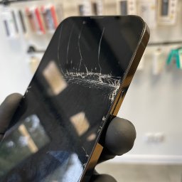 iBroken - Serwis Apple Kraków - Smartfon z pękniętym ekranem, trzymany w dłoni w czarnej rękawiczce, na tle regału z akcesoriami do telefonów.