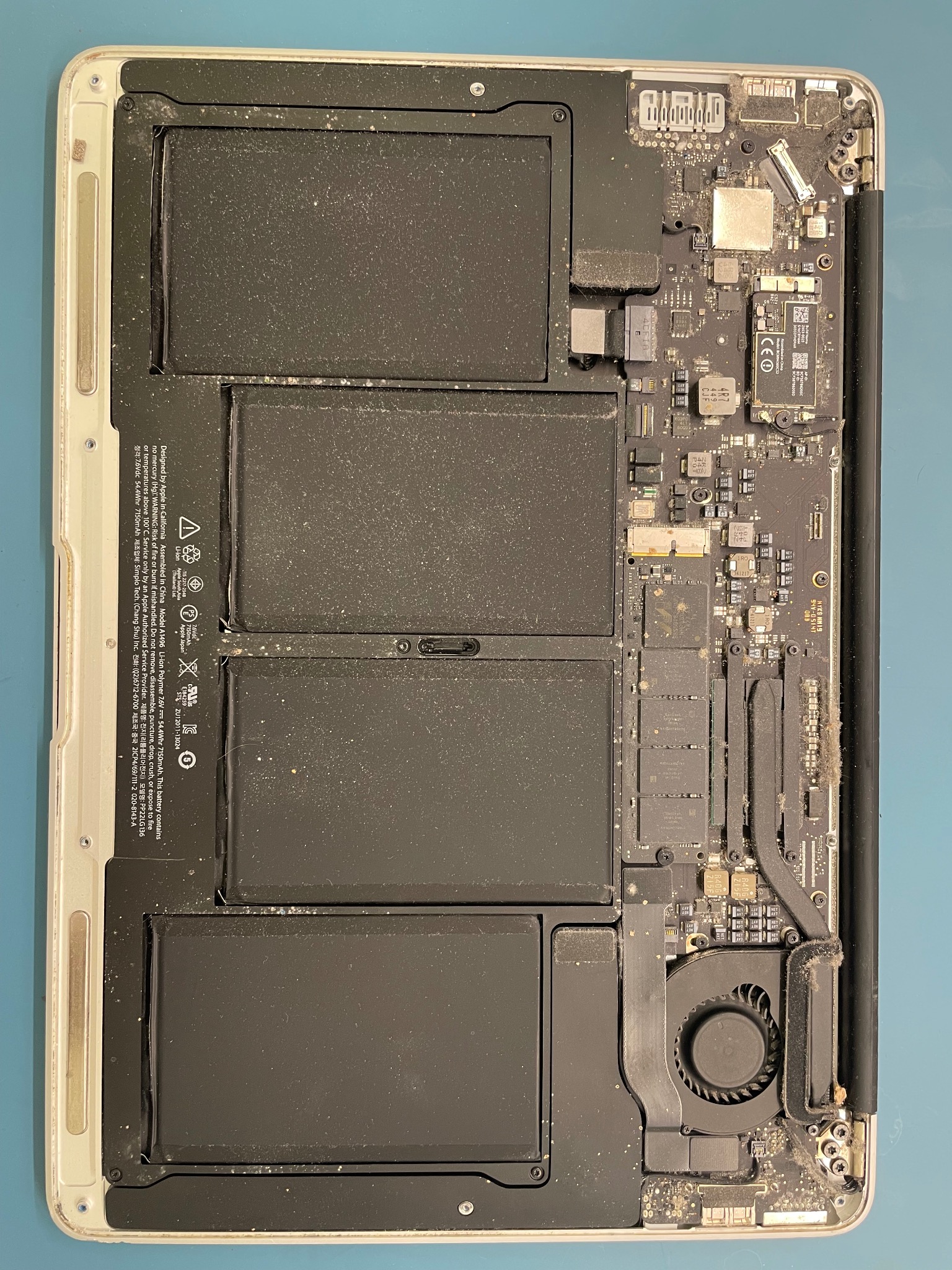 Wnętrze otwartego, zakurzonego laptopa Apple MacBook z widoczną baterią, płytą główną i wentylatorem chłodzącym.