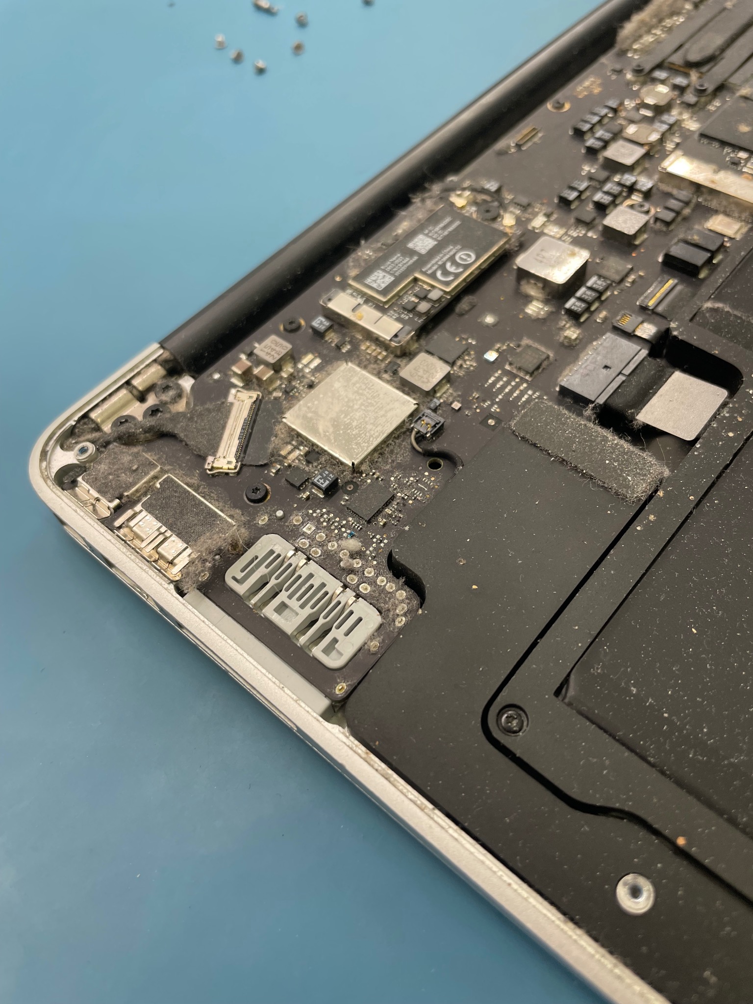 Wnętrze rozłożonego laptopa Apple MacBook Air z widoczną płytą główną, zanieczyszczeniami i odłączonymi elementami, na niebieskim tle warsztatowym.