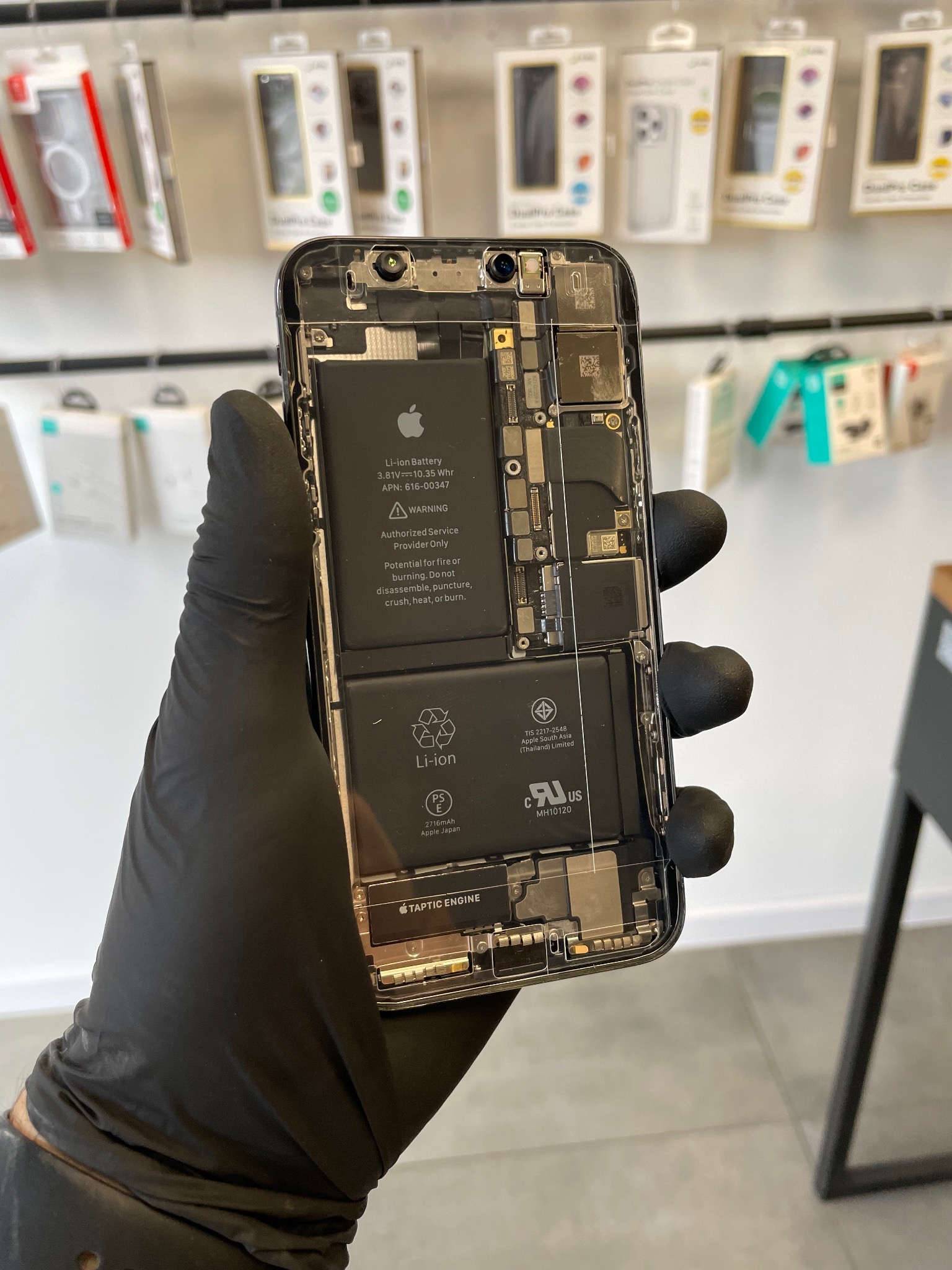 Otwarty telefon iPhone w rękawicy ochronnej, widoczne wewnętrzne komponenty, bateria i układ Taptic Engine, na tle półki z opakowaniami akcesoriów.