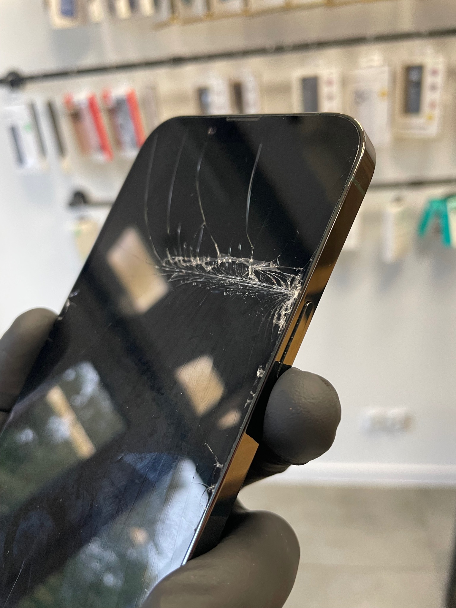 Smartfon z pękniętym ekranem, trzymany w dłoni w czarnej rękawiczce, na tle regału z akcesoriami do telefonów.
