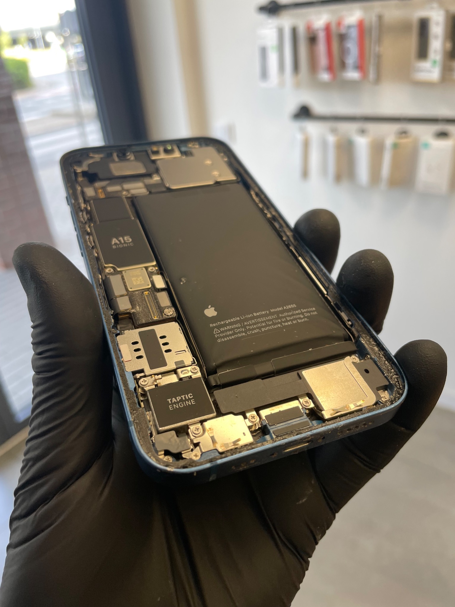 Rozłożony iPhone w rękawicy ochronnej, widoczne wnętrze z baterią, chipem A15 Bionic i Taptic Engine, na tle ściany z akcesoriami telefonicznymi.