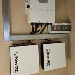 E-DYSTRYBUCJA SPÓŁKA Z OGRANICZONĄ ODPOWIEDZIALNOŚCIĄ - Kompletna instalacja: falownik Deye, dwa akumulatory Kon-Tec i zabezpieczenia. Elementy zamontowane na szynie, widoczne przewody. Całość na tle beżowej ściany.
