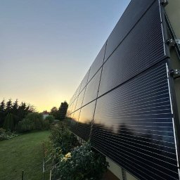 E-DYSTRYBUCJA SPÓŁKA Z OGRANICZONĄ ODPOWIEDZIALNOŚCIĄ - Panele fotowoltaiczne zamontowane na fasadzie budynku, odbijające zachód słońca. Widok z dołu, zieleń w tle.