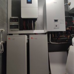 E-DYSTRYBUCJA SPÓŁKA Z OGRANICZONĄ ODPOWIEDZIALNOŚCIĄ - Kompletna instalacja z dwoma magazynami energii Felicity Solar, inwerterem i rozdzielnicą w pomieszczeniu z szarą płytką. Widoczny piec Panasonic.
