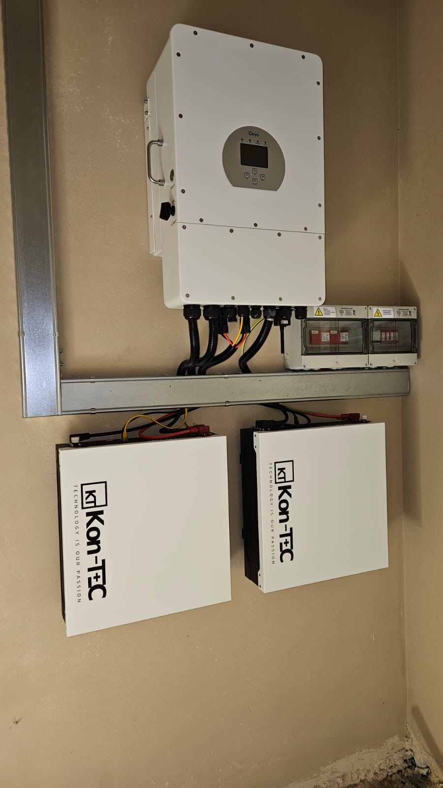 Kompletna instalacja: falownik Deye, dwa akumulatory Kon-Tec i zabezpieczenia. Elementy zamontowane na szynie, widoczne przewody. Całość na tle beżowej ściany.