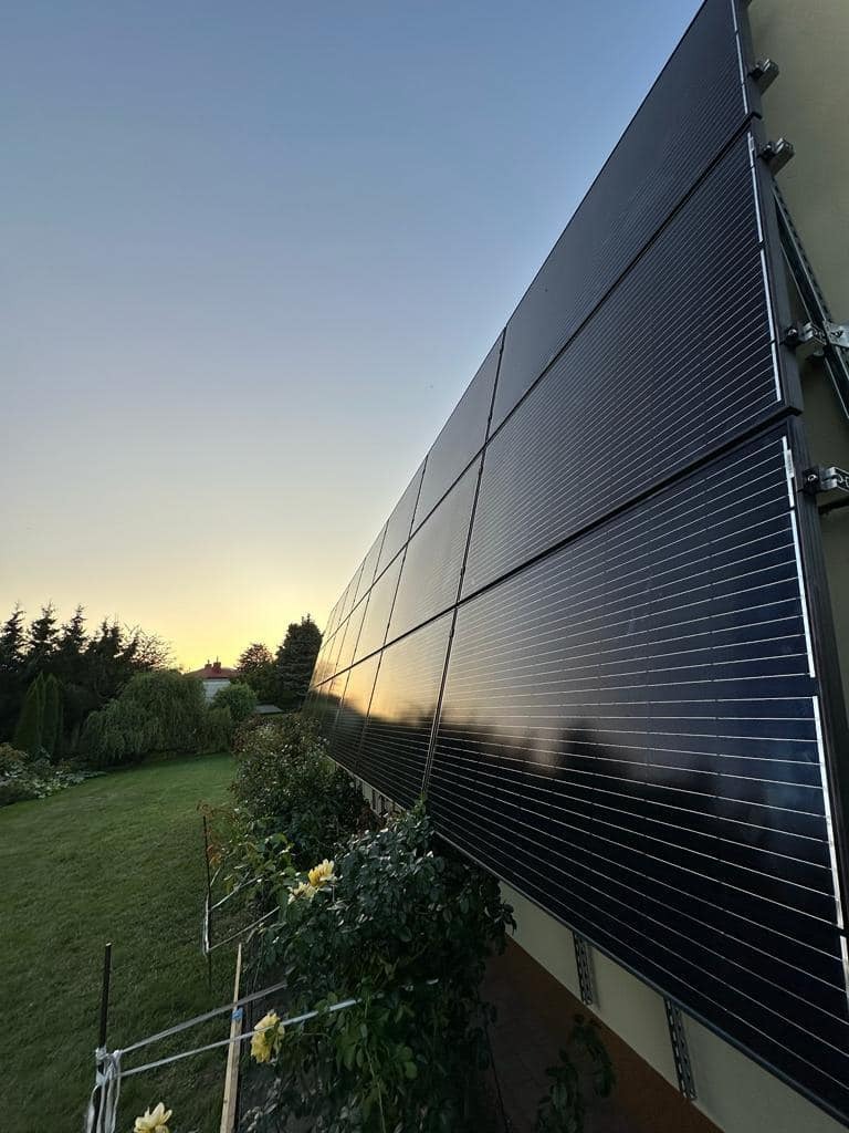 Panele fotowoltaiczne zamontowane na fasadzie budynku, odbijające zachód słońca. Widok z dołu, zieleń w tle.