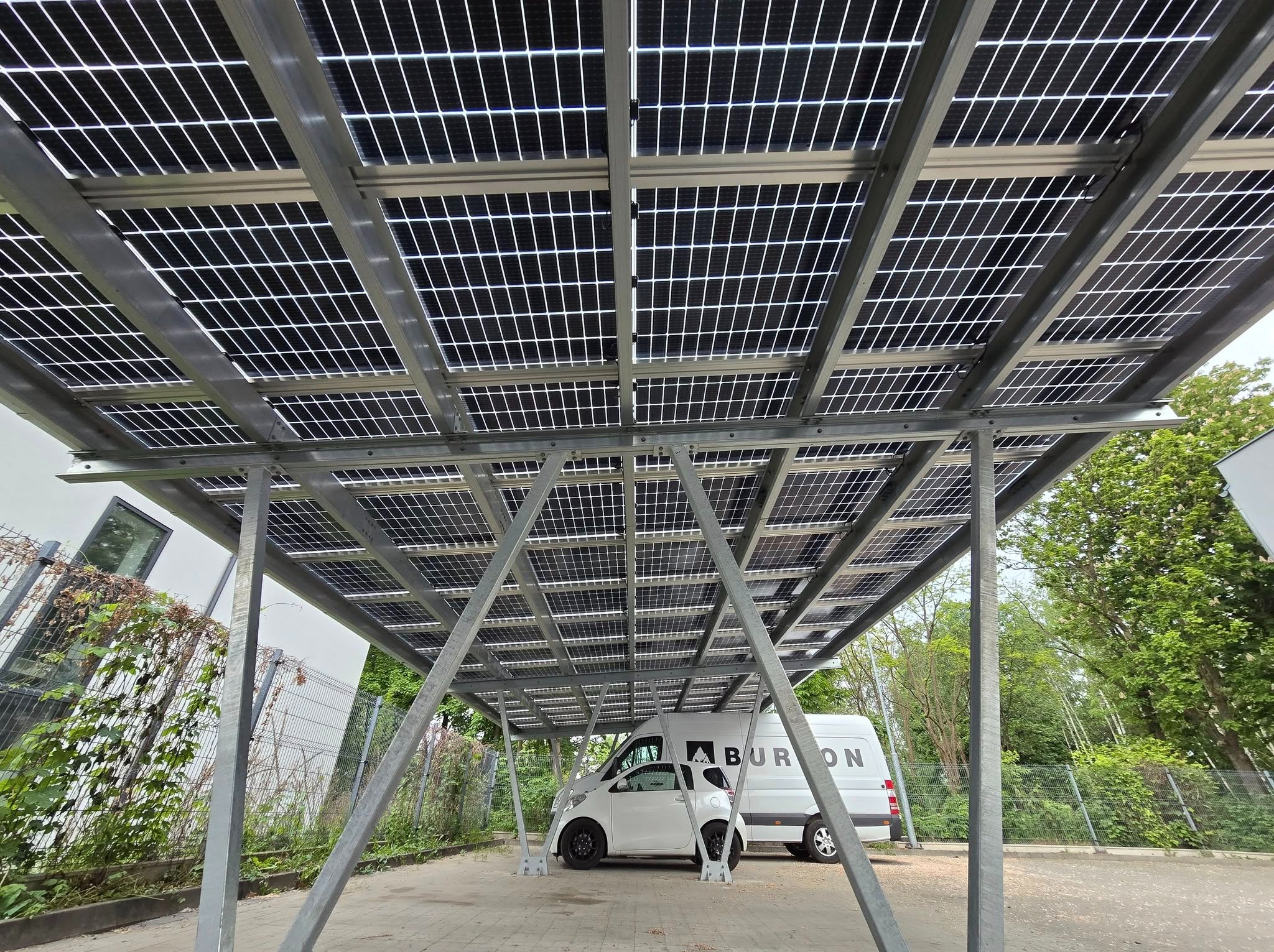 Zadaszenie z paneli fotowoltaicznych nad miejscami parkingowymi, wsparte na metalowej konstrukcji. W tle zaparkowane pojazdy.