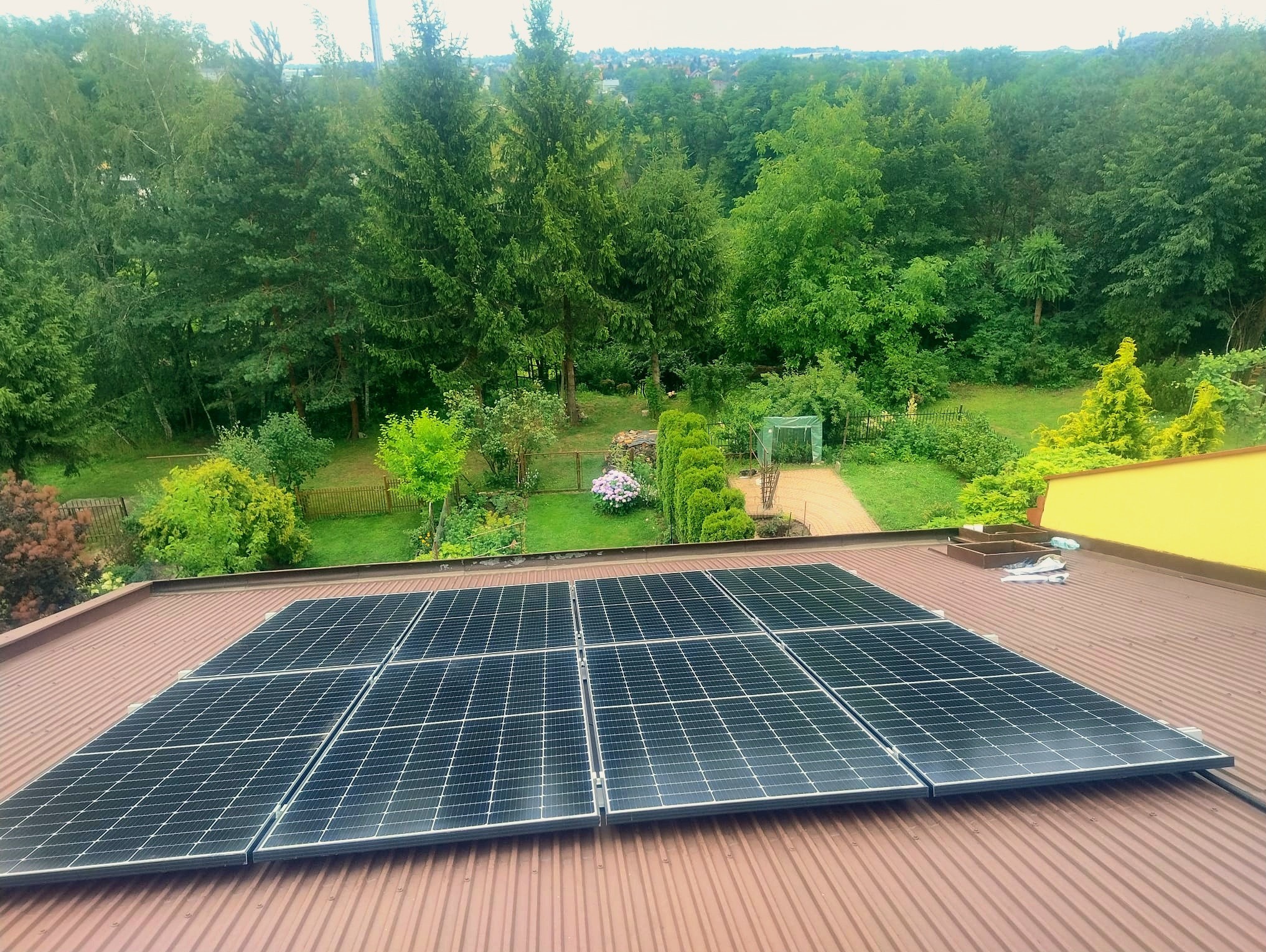 Panele słoneczne na brązowym dachu z blachy falistej, widok z góry na zieleń drzew i zabudowania. Część paneli poza kadrem.