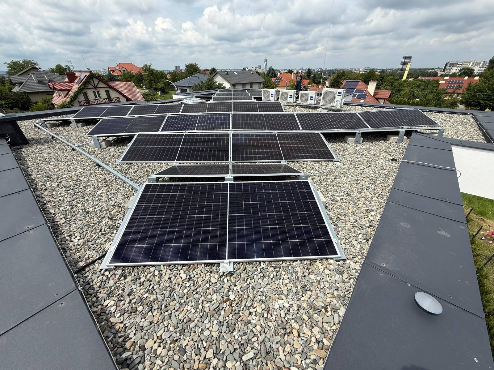 Instalacja fotowoltaiczna na dachu pokrytym kamieniami, widok z góry na panele, w tle zabudowa miejska i klimatyzatory. Montaż paneli słonecznych.