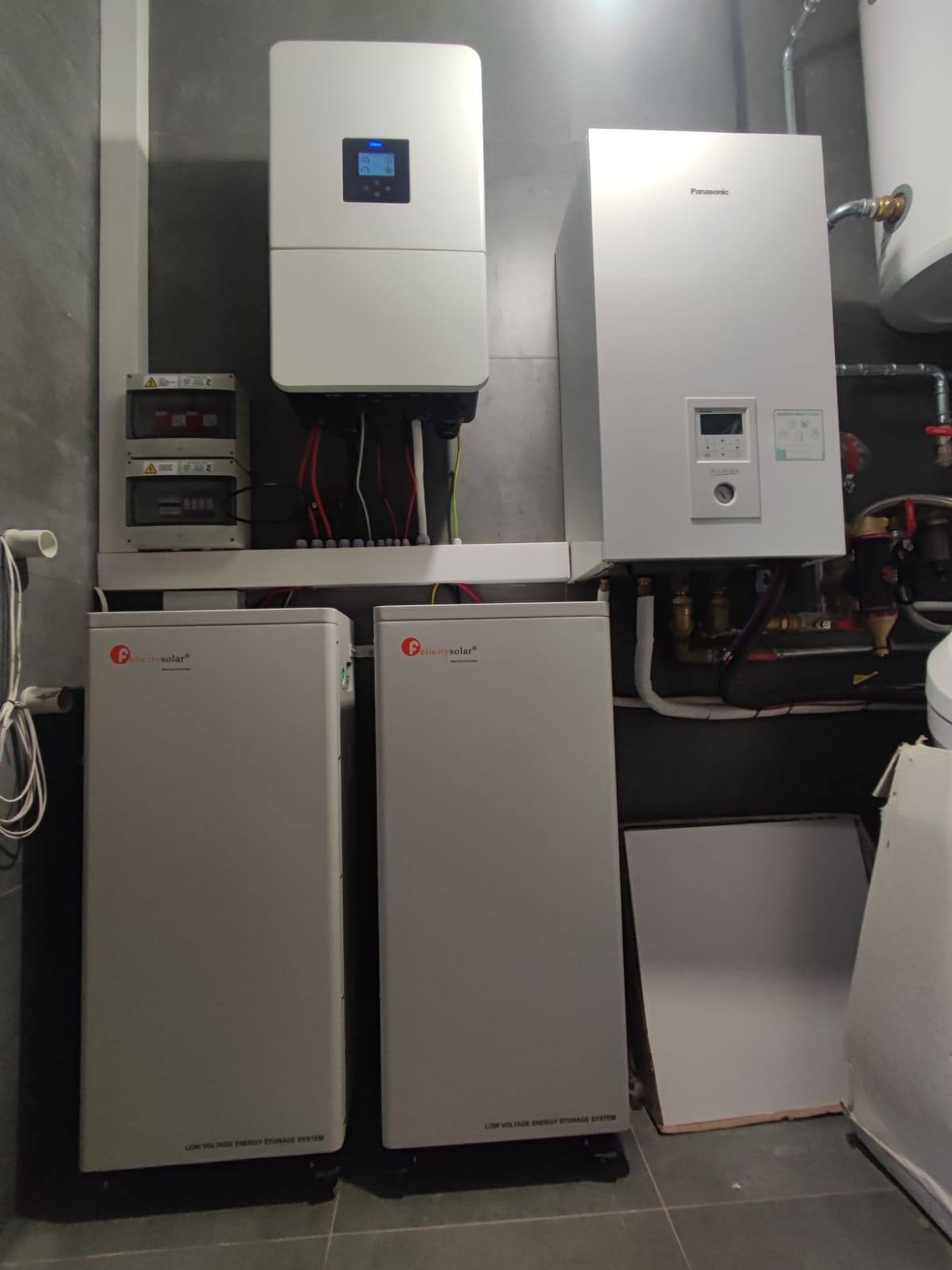 Kompletna instalacja z dwoma magazynami energii Felicity Solar, inwerterem i rozdzielnicą w pomieszczeniu z szarą płytką. Widoczny piec Panasonic.