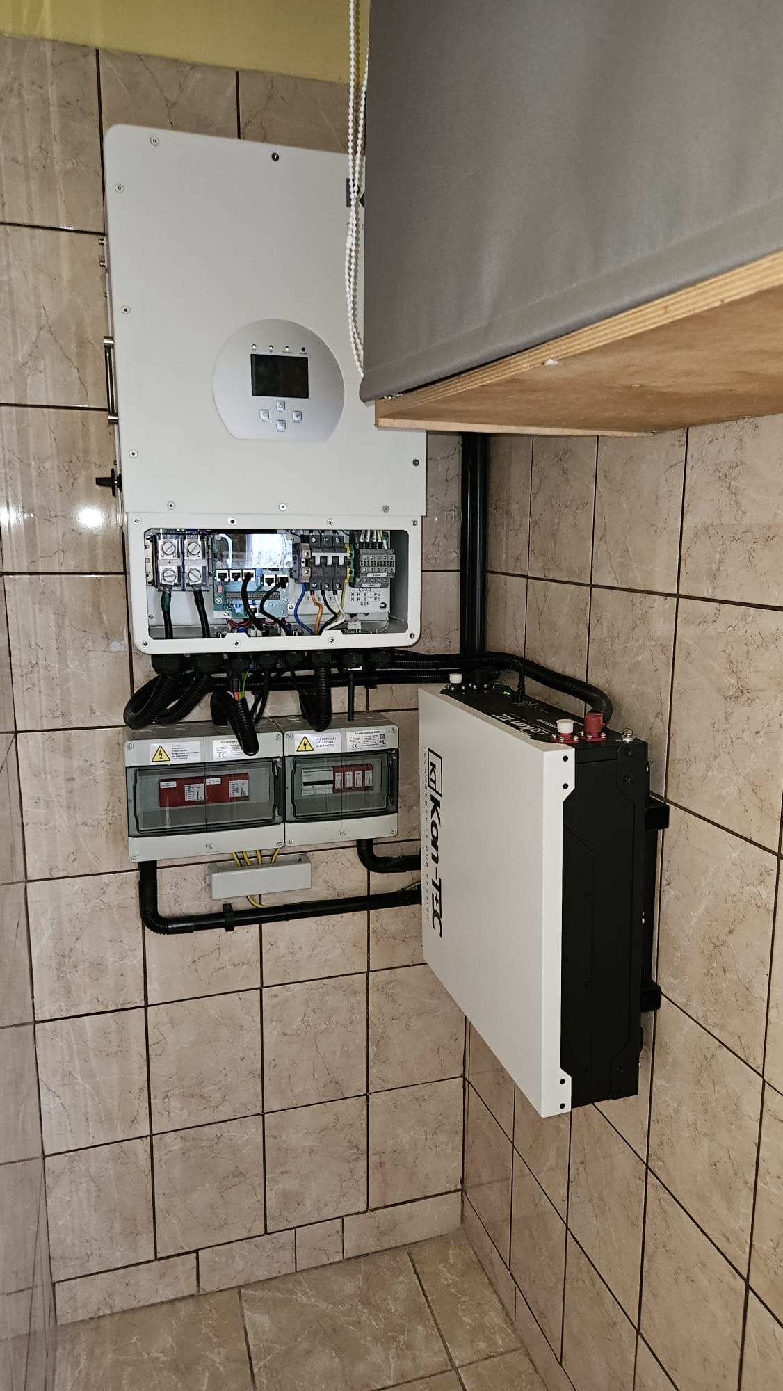Kompletna instalacja magazynu energii Kon-Tec na ścianie z beżowych płytek, widoczne zabezpieczenia i okablowanie, roleta okienna w tle.