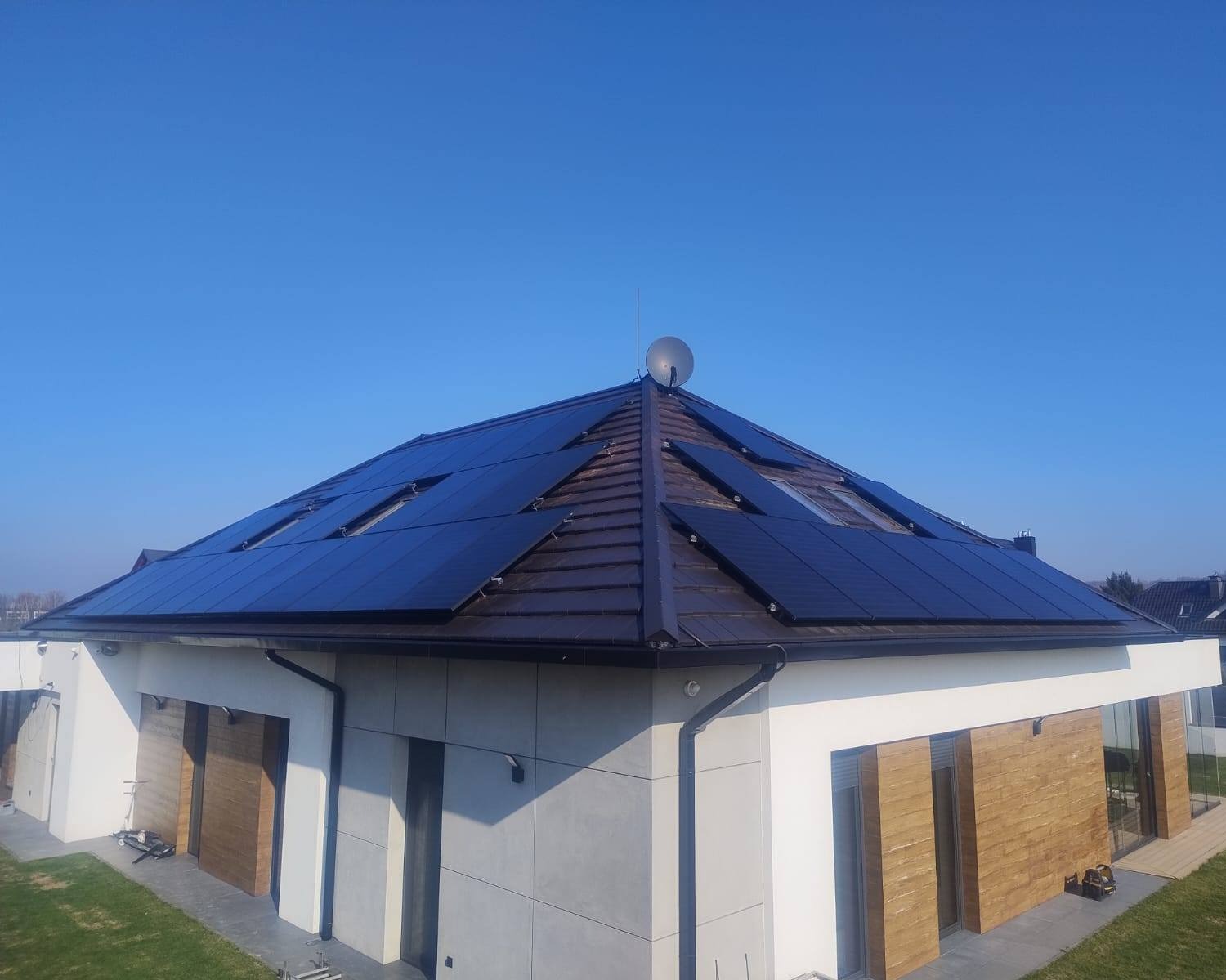 Panele słoneczne na brązowym dachu domu jednorodzinnego z jasną elewacją i anteną satelitarną na szczycie, błękitne niebo w tle.