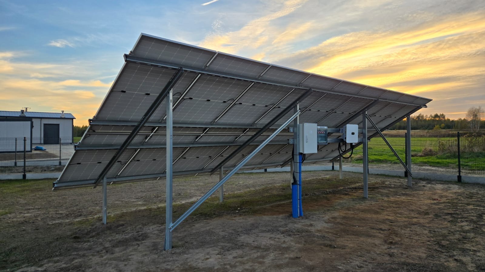 Widok od spodu na panele fotowoltaiczne zamontowane na metalowej konstrukcji. W tle budynek i zachodzące słońce.