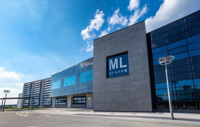 Budynek biurowy z panelami fotowoltaicznymi na fasadzie, logo ML System w oknie, błękitne niebo z chmurami w tle.