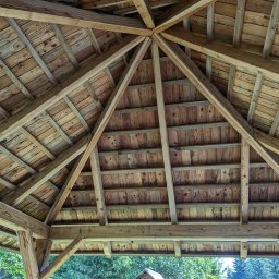 PPUH Pierlak szymon - Drewniana konstrukcja dachu altany z widocznymi belkami nośnymi i deskami, z zewnątrz widok na zieleń drzew.