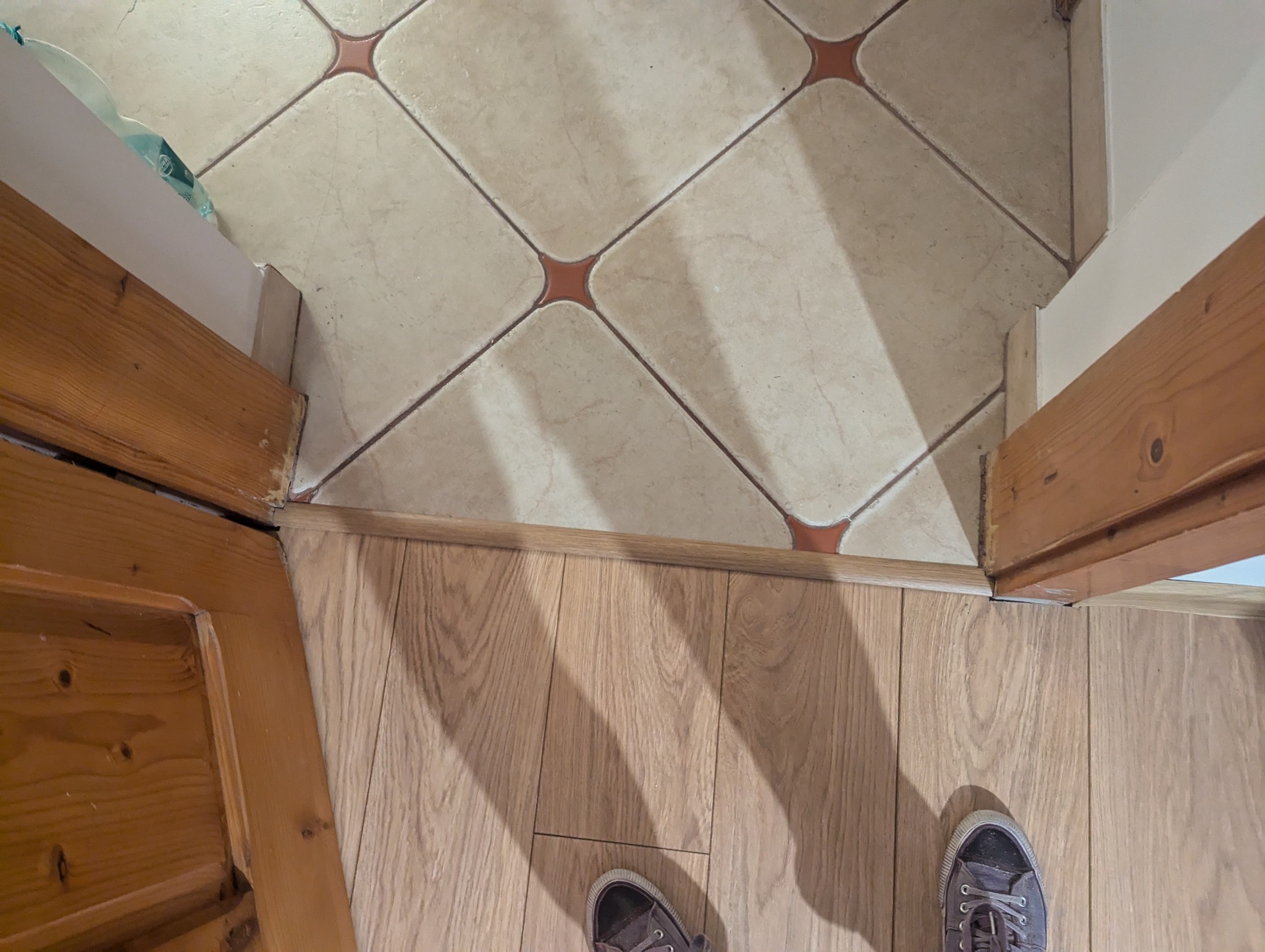 Połączenie płytek podłogowych z panelami podłogowymi przy futrynie drzwiowej, widoczne buty osoby robiącej zdjęcie.