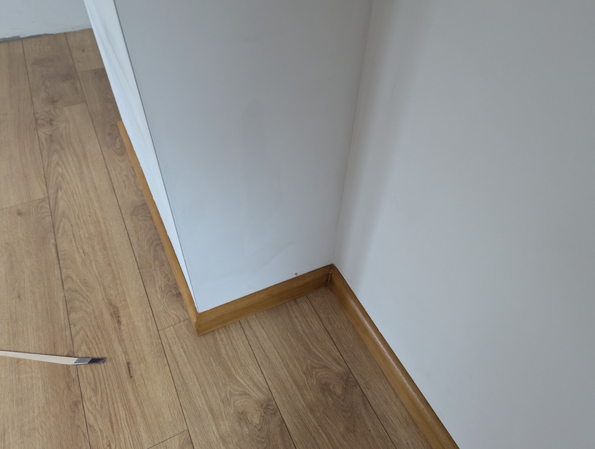 Szczegółowe ujęcie narożnika pokoju po remoncie: świeżo pomalowana biała ściana, drewniana listwa przypodłogowa i laminowana podłoga w odcieniach brązu, na podłodze leży pędzel.