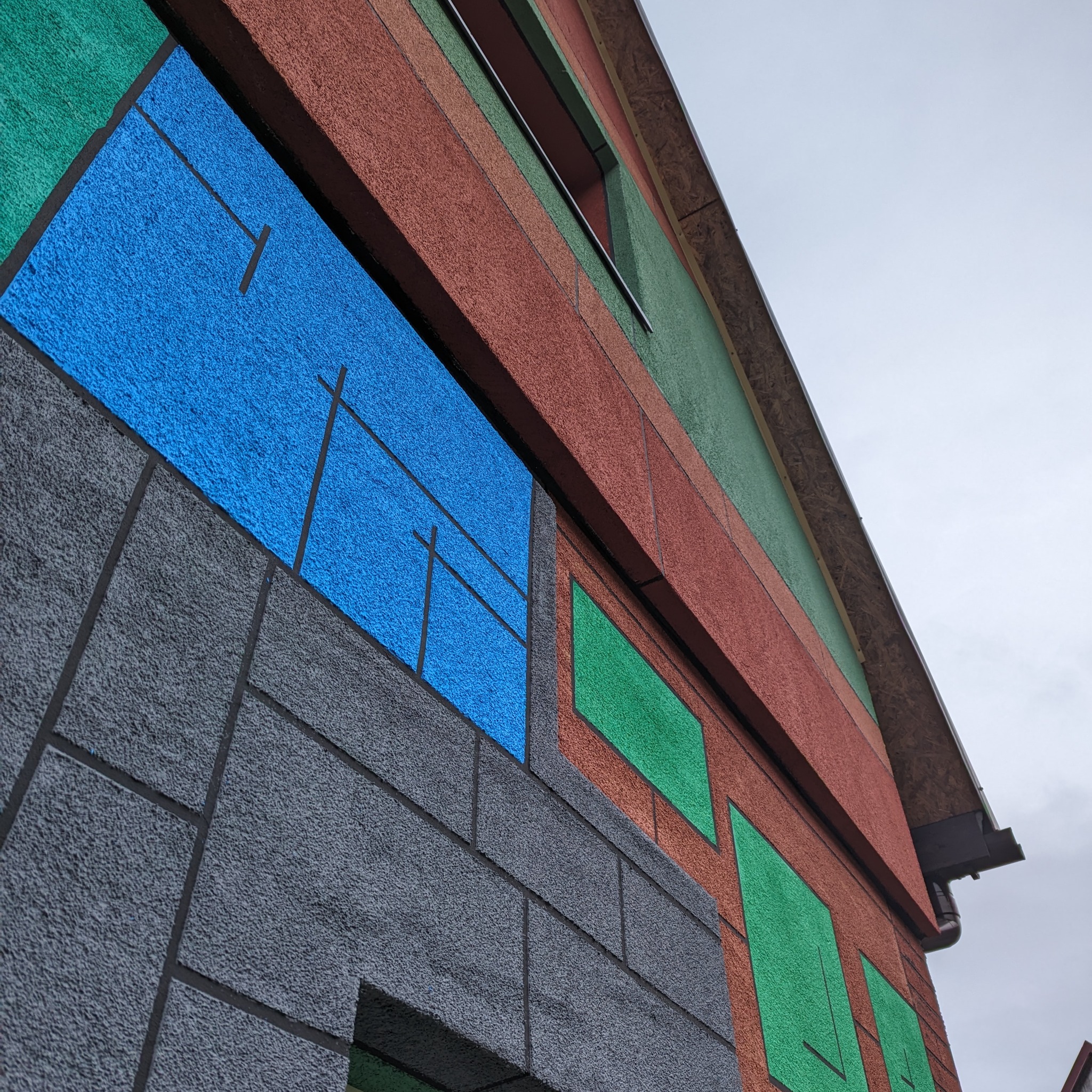 Kolorowa elewacja domu z tynkiem strukturalnym w odcieniach szarości, błękitu, zieleni i czerwieni, z widocznym oknem i fragmentem dachu z desek.