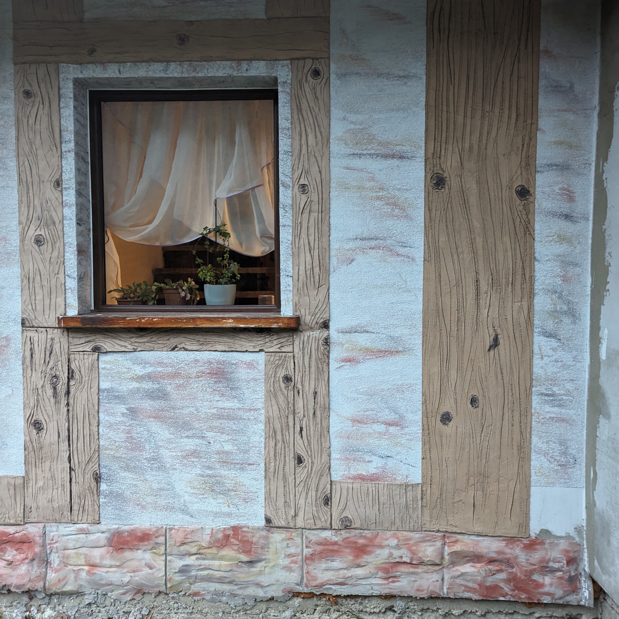 Fasada budynku z oknem i imitacją drewna, wykonana w jasnych kolorach z elementami dekoracyjnymi i roślinami w doniczkach na parapecie.