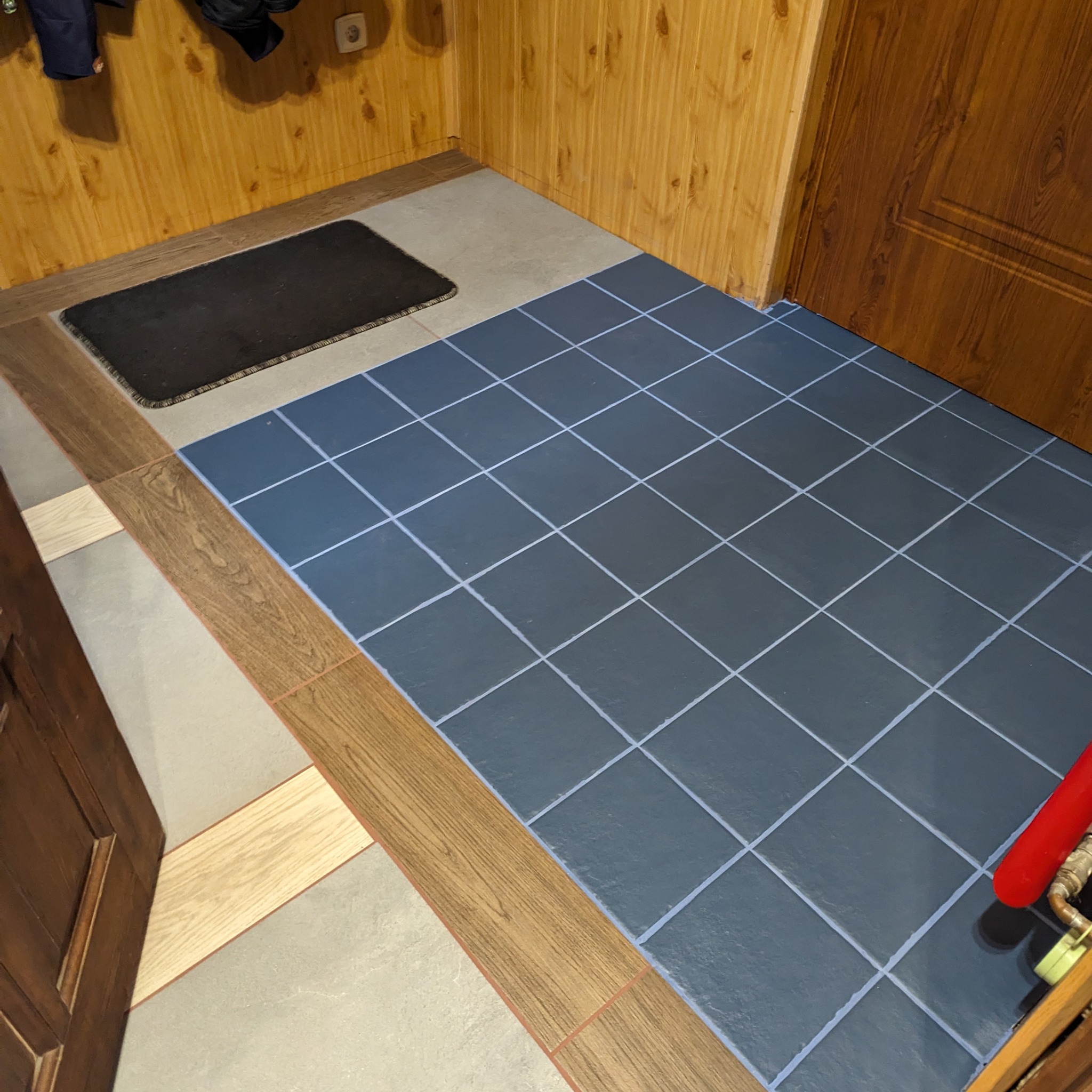 Wykończony fragment podłogi w przedpokoju z ciemnymi płytkami ułożonymi w kwadratową siatkę z jasnymi fugami, listwami przypodłogowymi imitującymi drewno oraz fragmentem drewnianej ściany i drzwi.