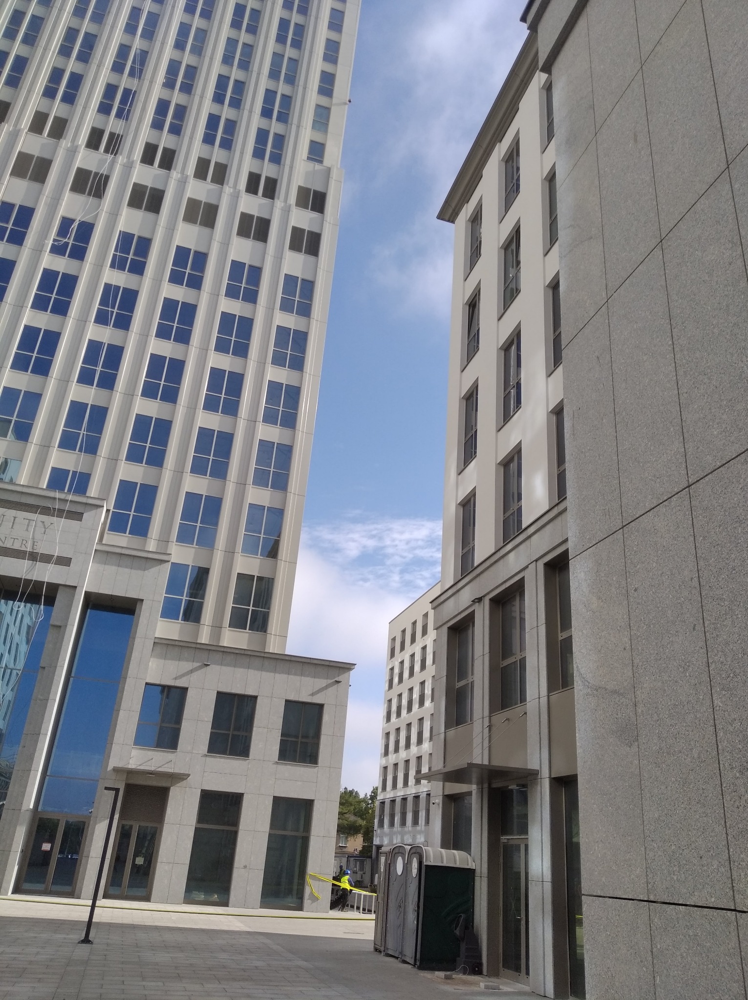 Dwa budynki biurowe o nowoczesnej architekturze, z widocznym fragmentem nieba, oraz przenośne toalety na chodniku.