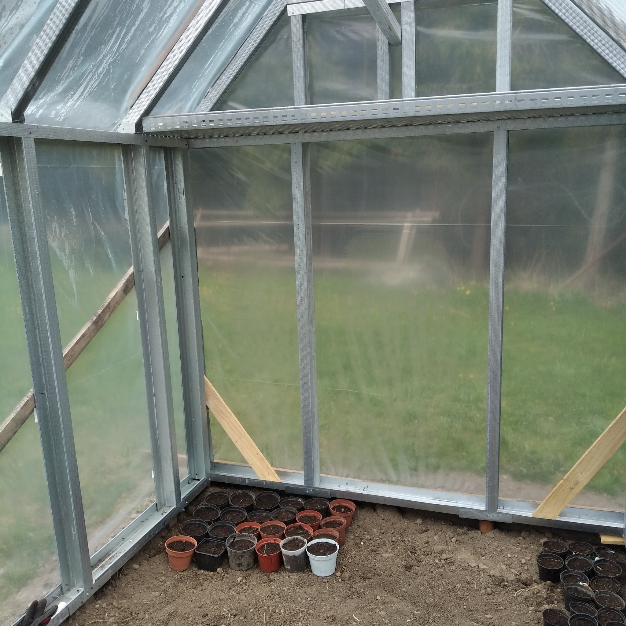 Wnętrze szklarni z metalową konstrukcją i przezroczystym pokryciem, z widocznymi doniczkami z ziemią przygotowanymi do sadzenia roślin.
