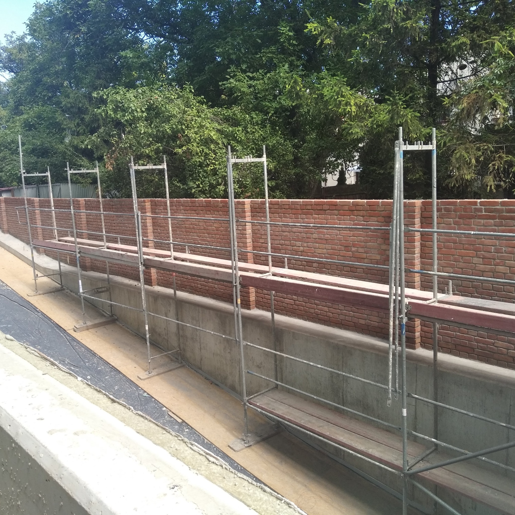 Murowana ściana z czerwonej cegły w trakcie budowy, widoczna ze rusztowania na tle zielonych drzew.