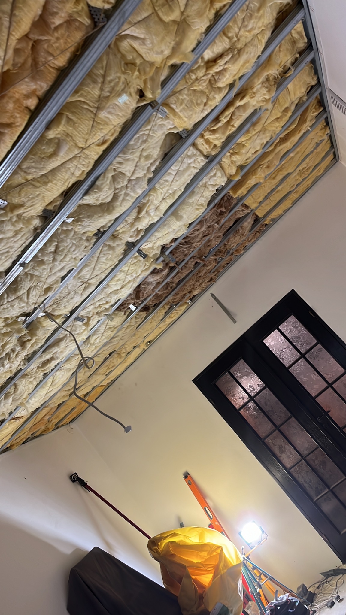 Montaż izolacji z wełny mineralnej pomiędzy metalowymi profilami sufitu podwieszanego, widoczne fragmenty niezabezpieczonej izolacji, okno w tle, narzędzia i oświetlenie robocze