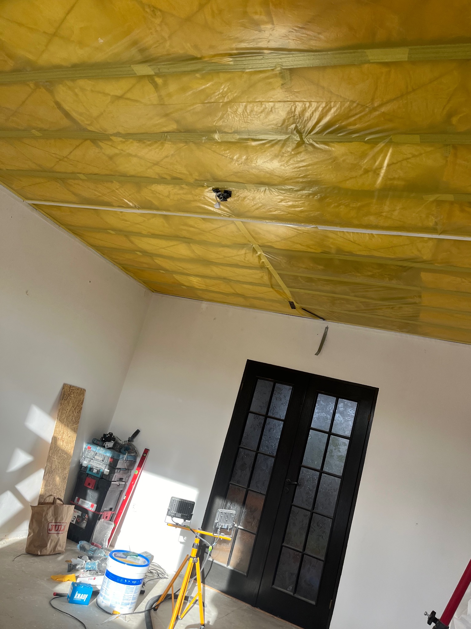 Żółta folia ochronna zabezpiecza sufit z zamontowaną listwą, przygotowany do dalszych prac wykończeniowych. W tle widoczne czarne, przeszklone drzwi oraz narzędzia i materiały budowlane w rogu...