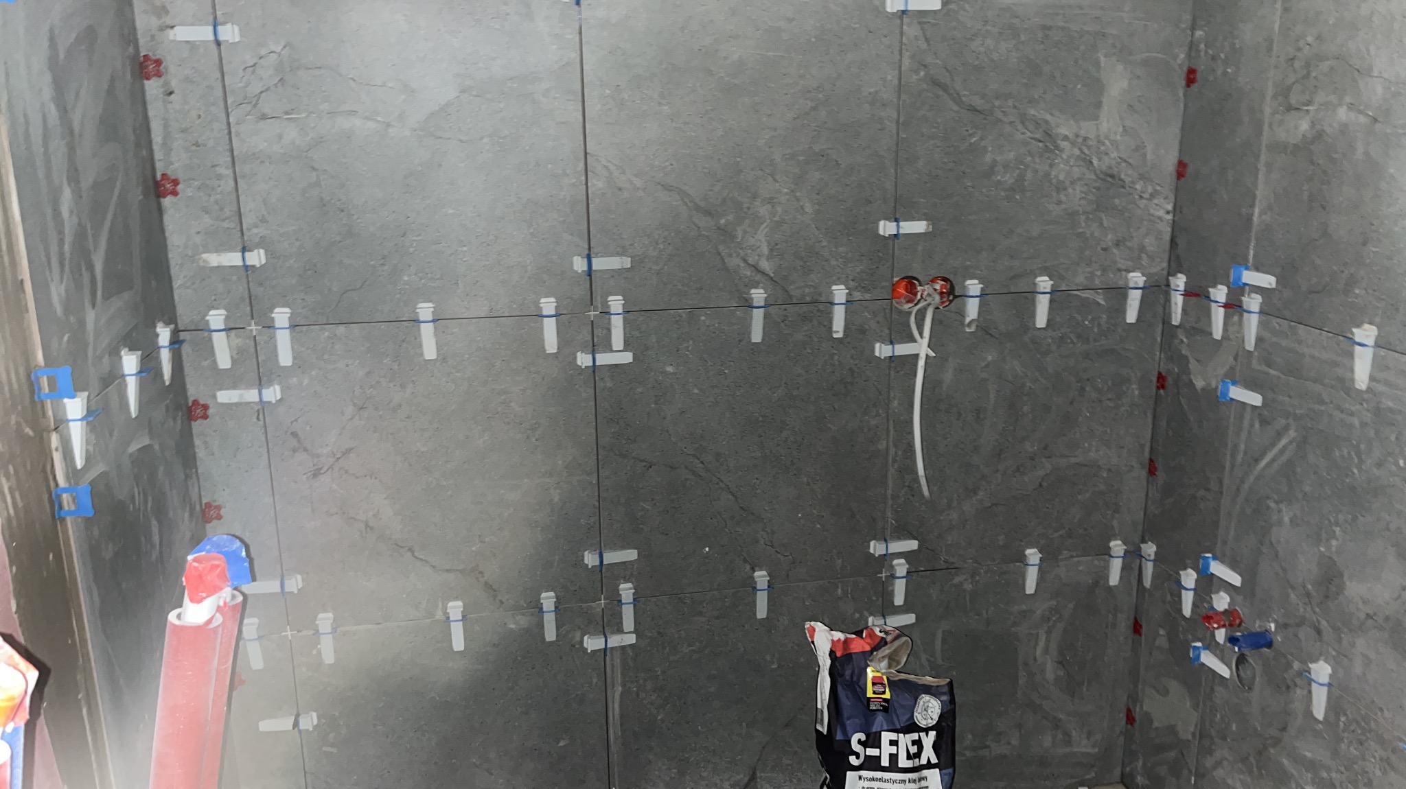 Ściana łazienki w trakcie układania szarych płytek z użyciem systemu poziomowania płytek, widoczne przewody elektryczne i worek z klejem elastycznym S-FLEX.