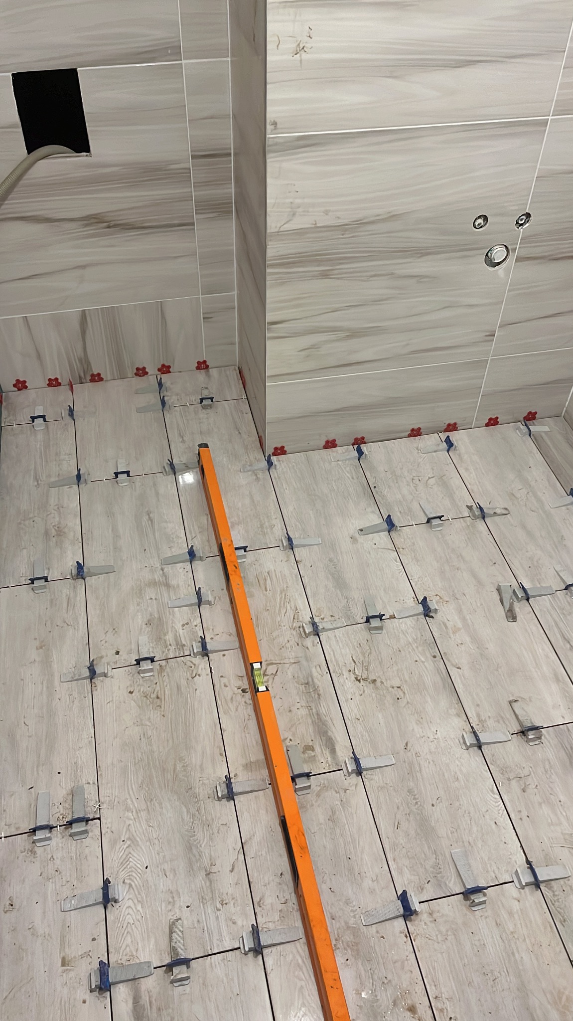 Układanie płytek podłogowych imitujących drewno w łazience, z użyciem poziomicy i systemu poziomowania płytek, widoczne otwory na rury na ścianie.