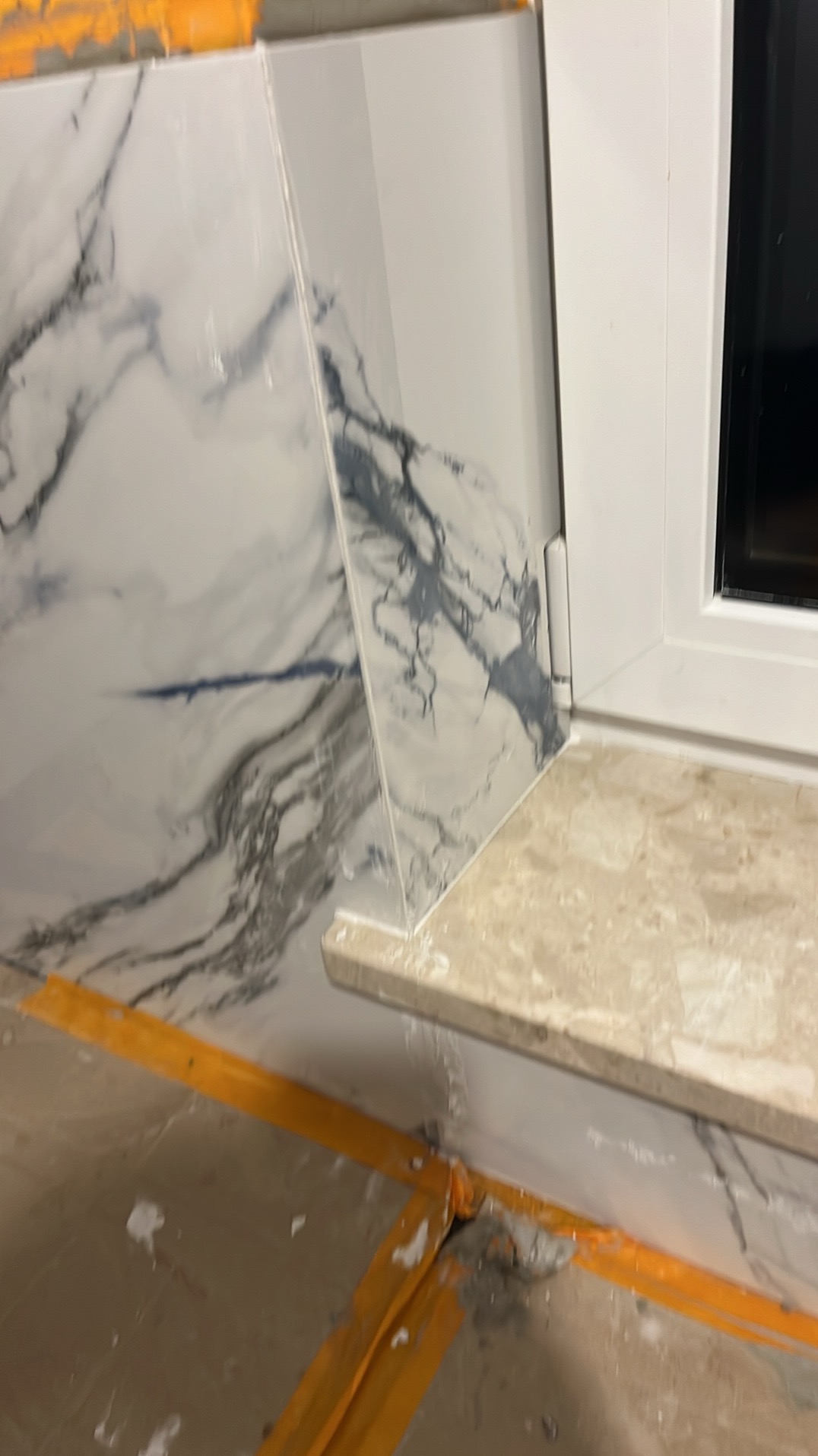 Fragment łazienki w trakcie wykańczania: ułożone płytki imitujące marmur na ścianie, zabezpieczone żółtą taśmą, widoczny parapet z jasnego kamienia przy oknie.