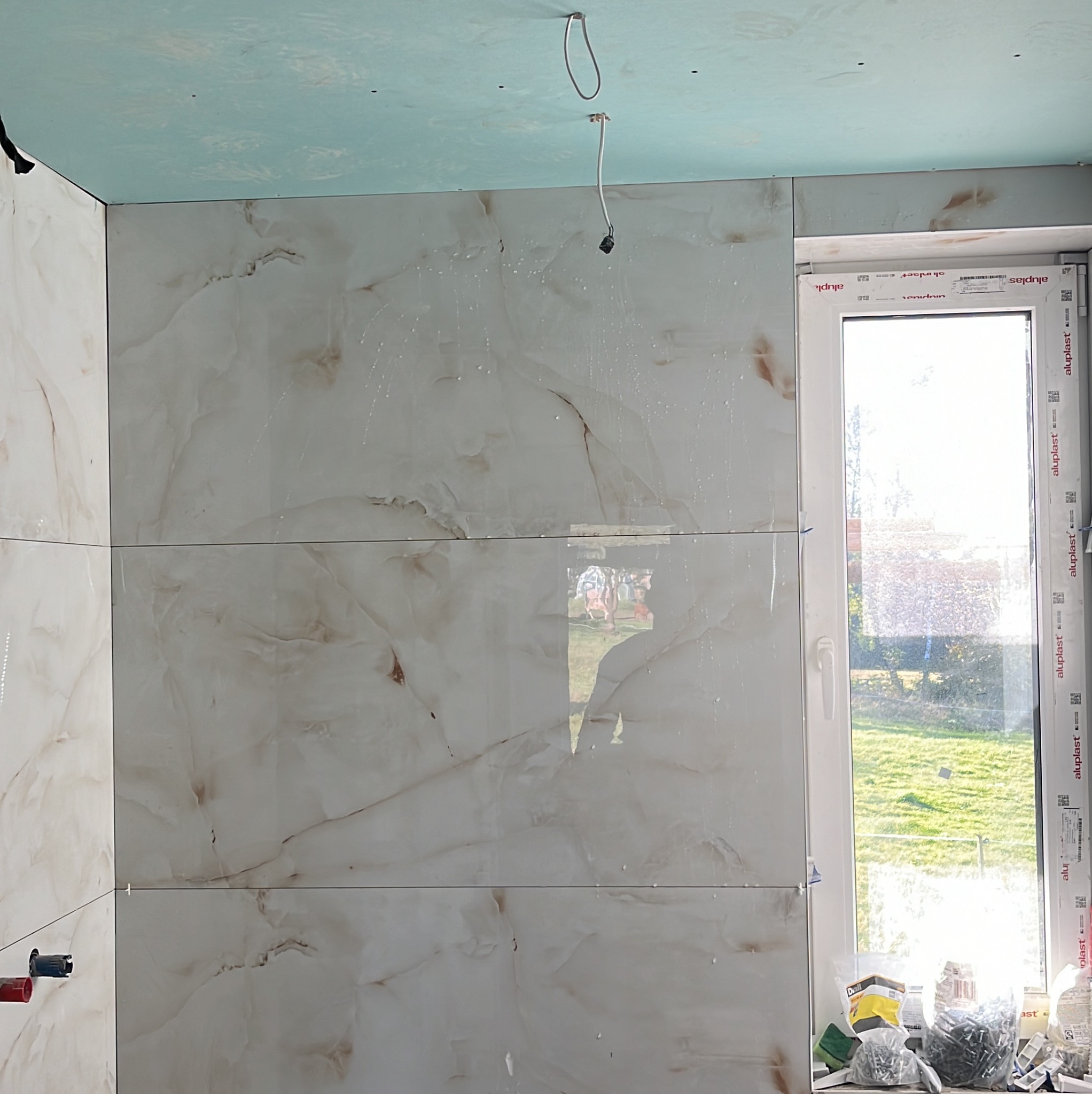 Montaż płytek imitujących marmur na ścianie łazienki, widoczne krzyżyki dystansowe, surowy sufit z płyt kartonowo-gipsowych i okno z widokiem na zewnątrz.