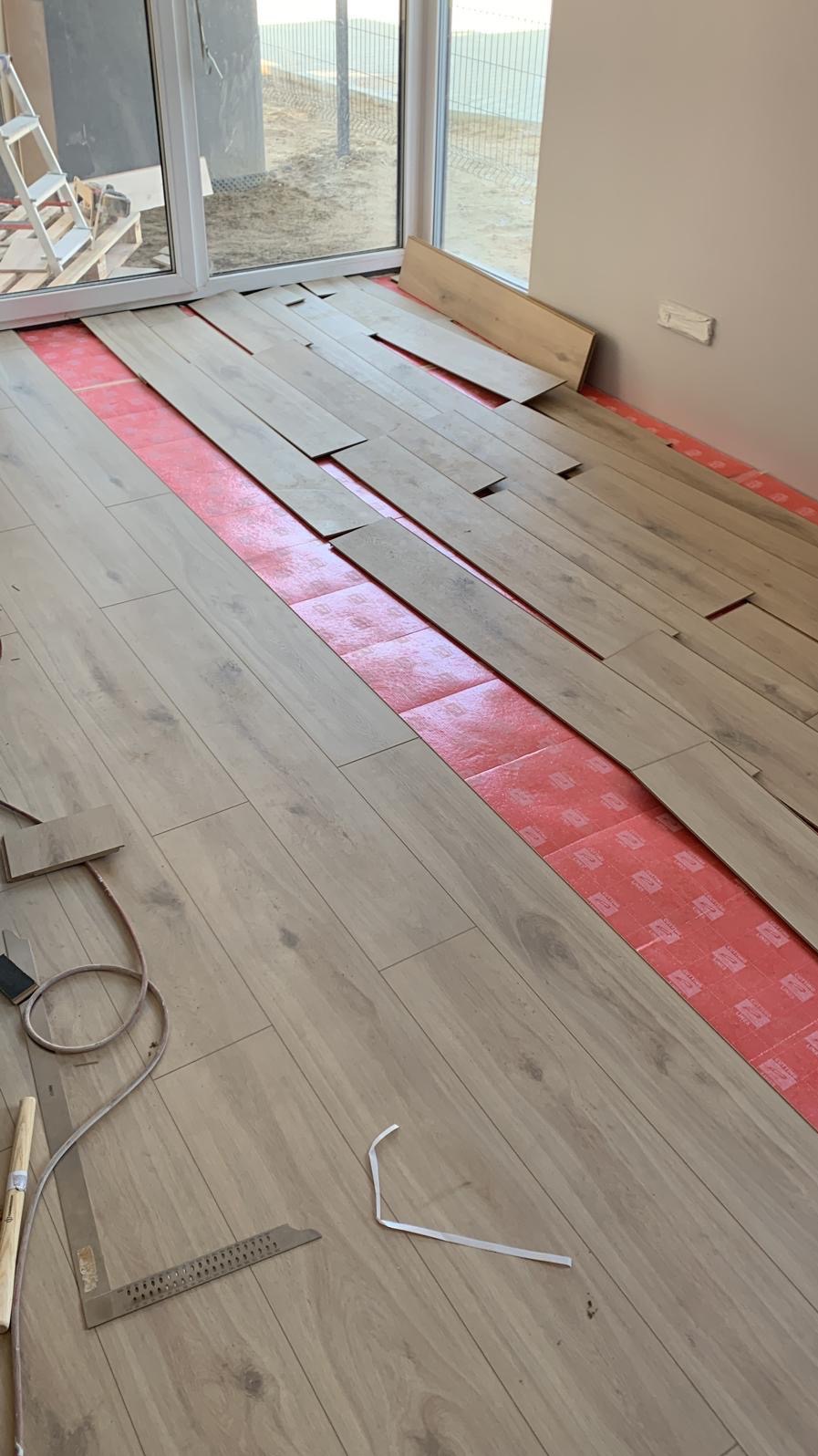 Układanie paneli podłogowych w jasnym odcieniu drewna na czerwonej folii izolacyjnej; widoczne narzędzia, kątownik i listwa przypodłogowa, duże okno balkonowe w tle.
