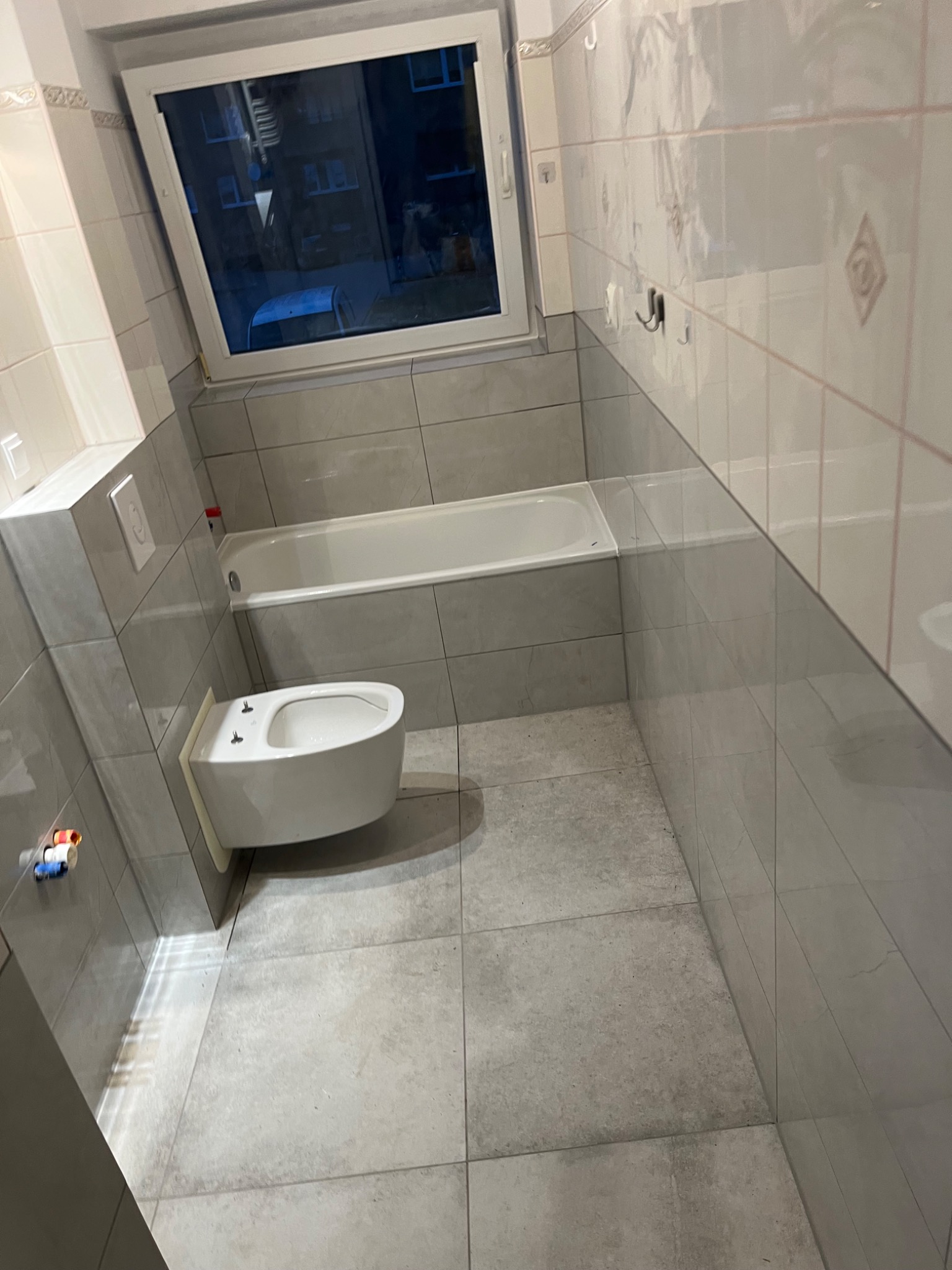 Wykończona łazienka z szarymi płytkami: podwieszany bidet, wanna, okno. Widoczne fugi i detale wykończeniowe.