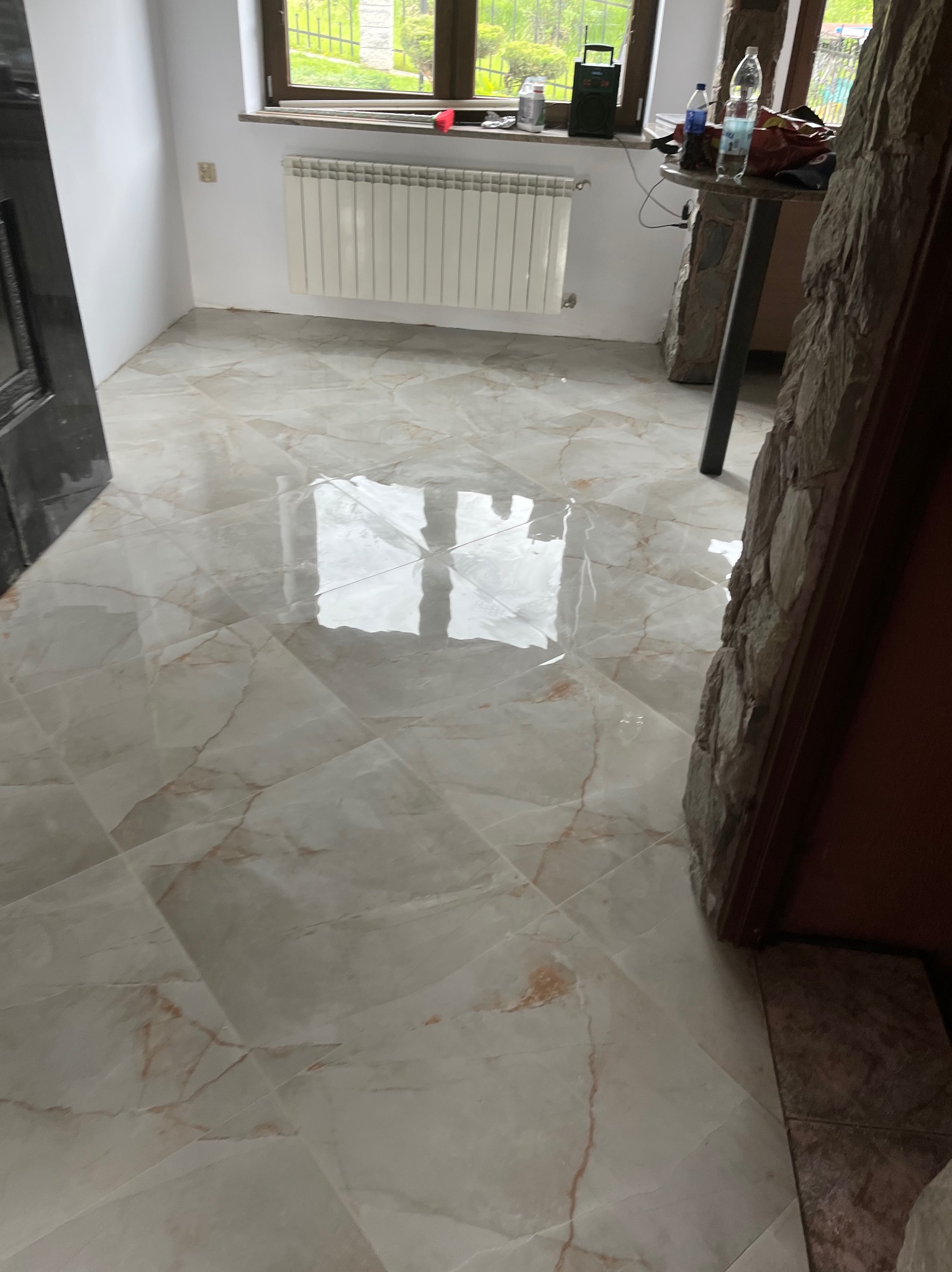 Położone, błyszczące płytki podłogowe w odcieniach beżu i brązu, z widocznym fragmentem okna i grzejnika w tle. Widoczny fragment kamiennej obudowy.