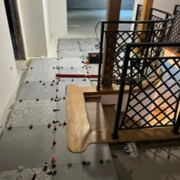 Układanie płytek podłogowych na schodach i korytarzu, widoczne poziomice, kliny dystansowe i wzorzyste płytki w szarościach, balustrada schodowa z metaloplastyki