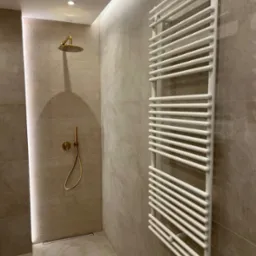 Nowoczesna kabina prysznicowa z złotą armaturą i białym grzejnikiem drabinkowym na beżowych płytkach.